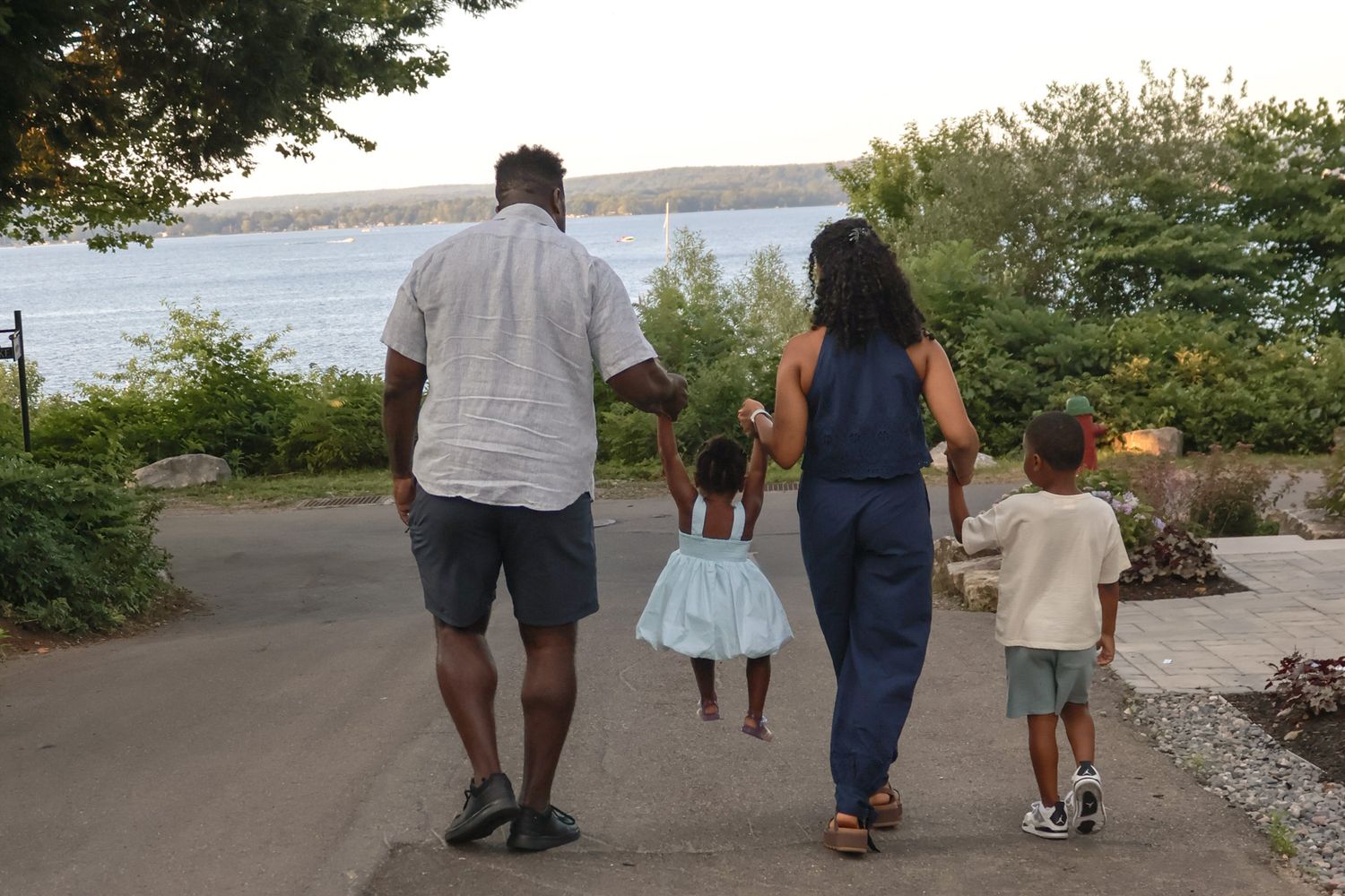 Family of four walking together along a waterfront pathway surrounded by lush green trees at dusk.