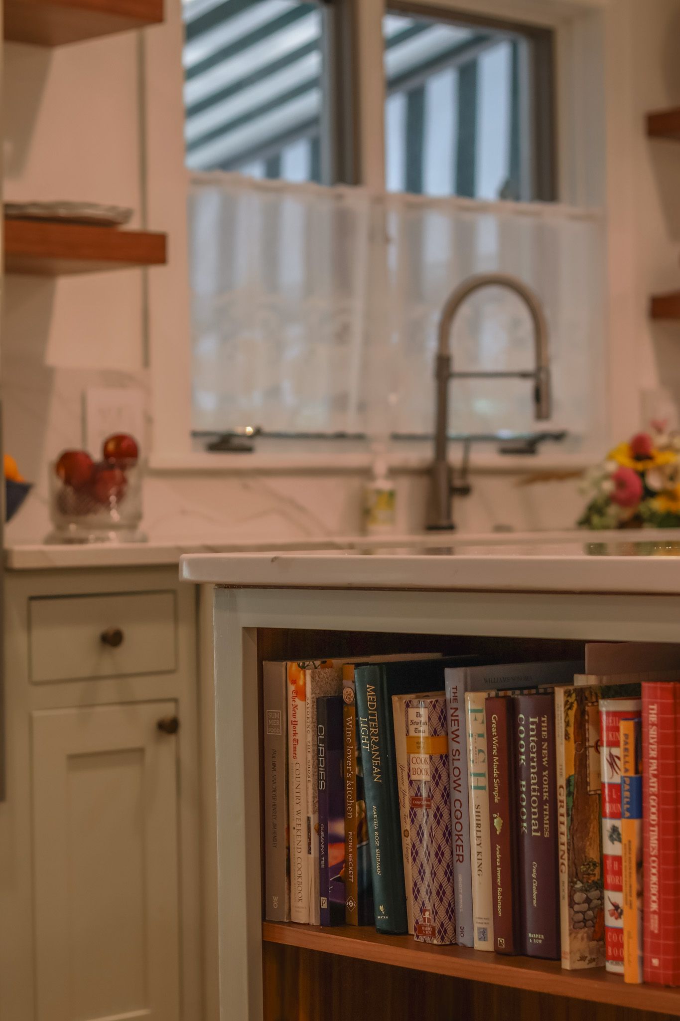 Cookbook shelf built into kitchen island with farmhouse sink and marble backsplash in background.