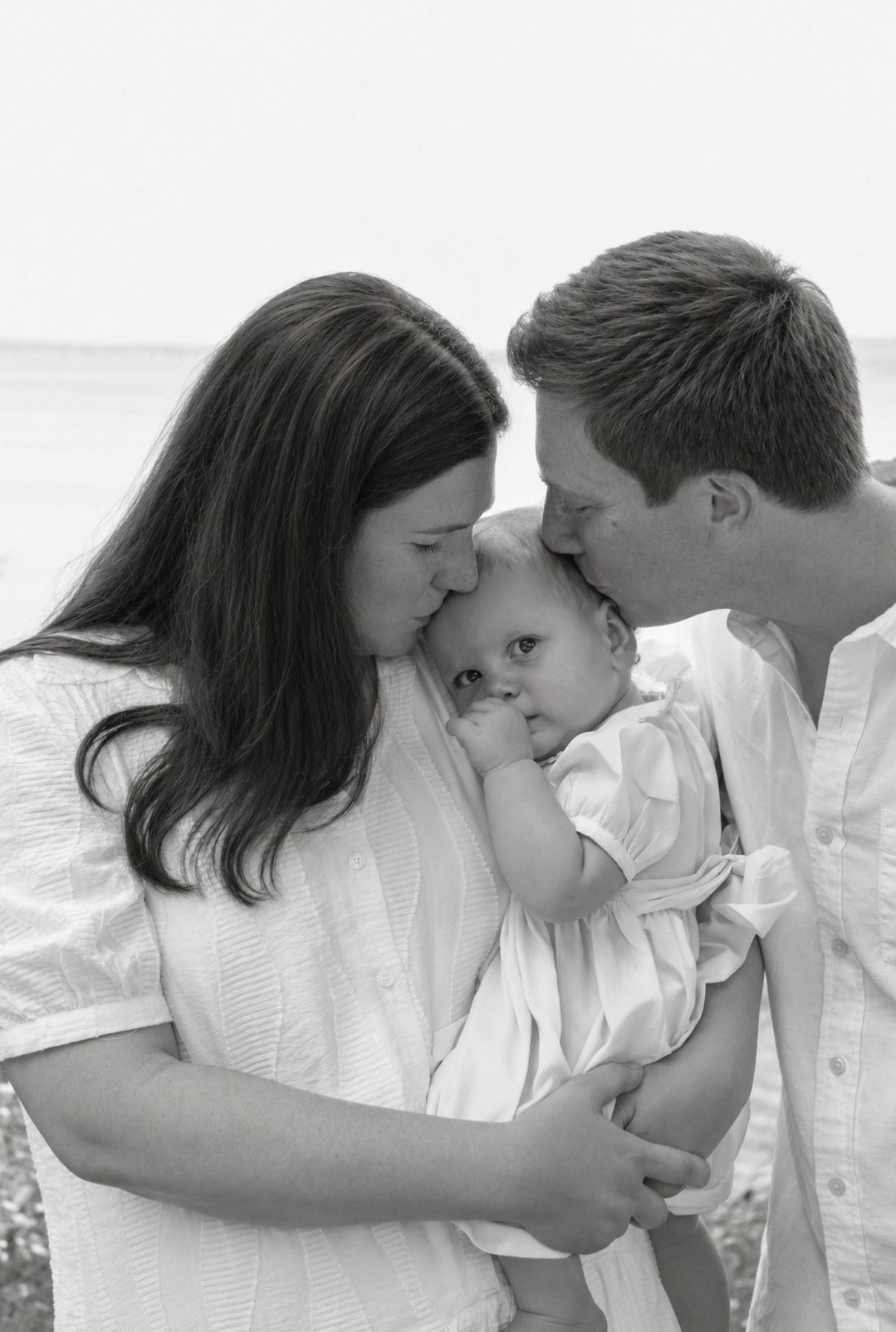 Black and white photo of parents kissing their baby on the forehead at a beach.