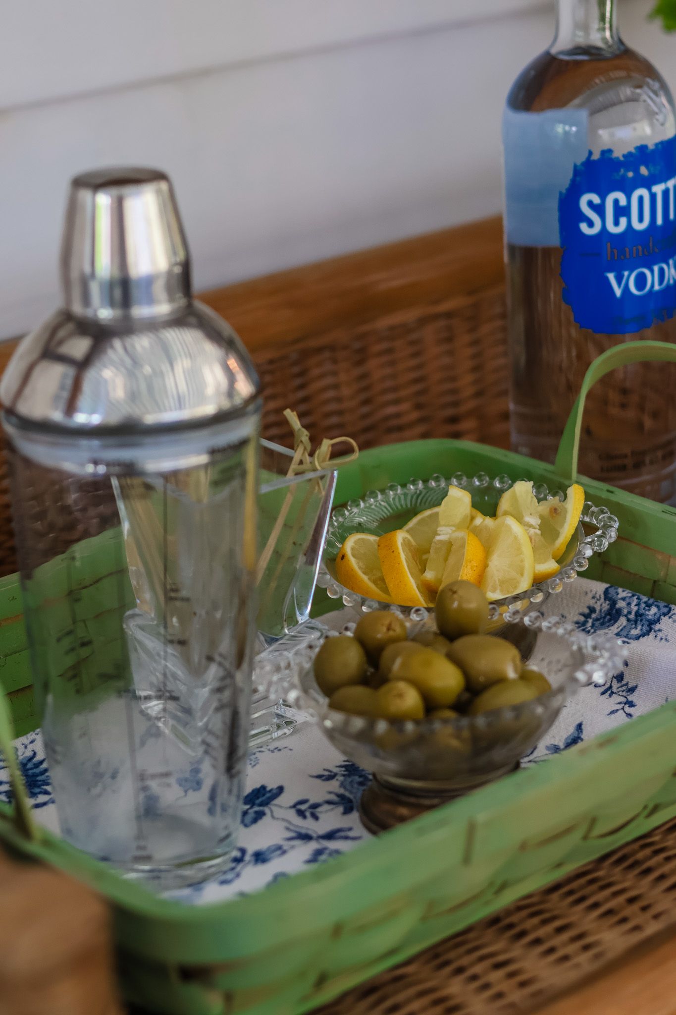 Cocktail tray with shaker, olives, lemon slices, and Scotch vodka bottle on wicker surface.
