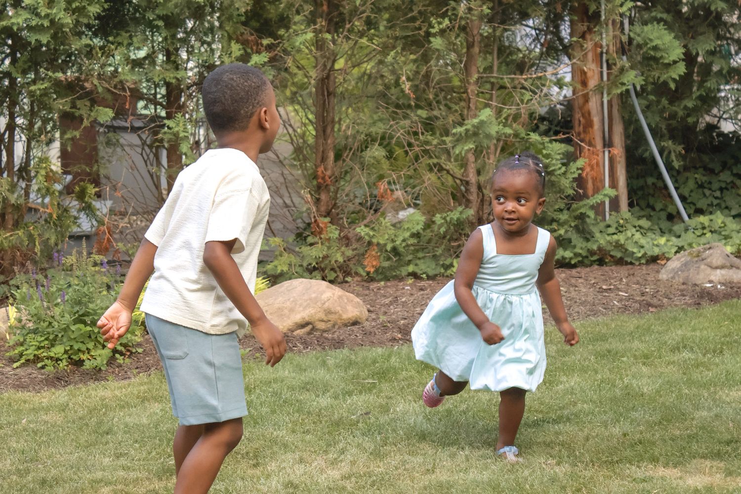 Two Black children playing and running together on a lush green lawn in a garden.