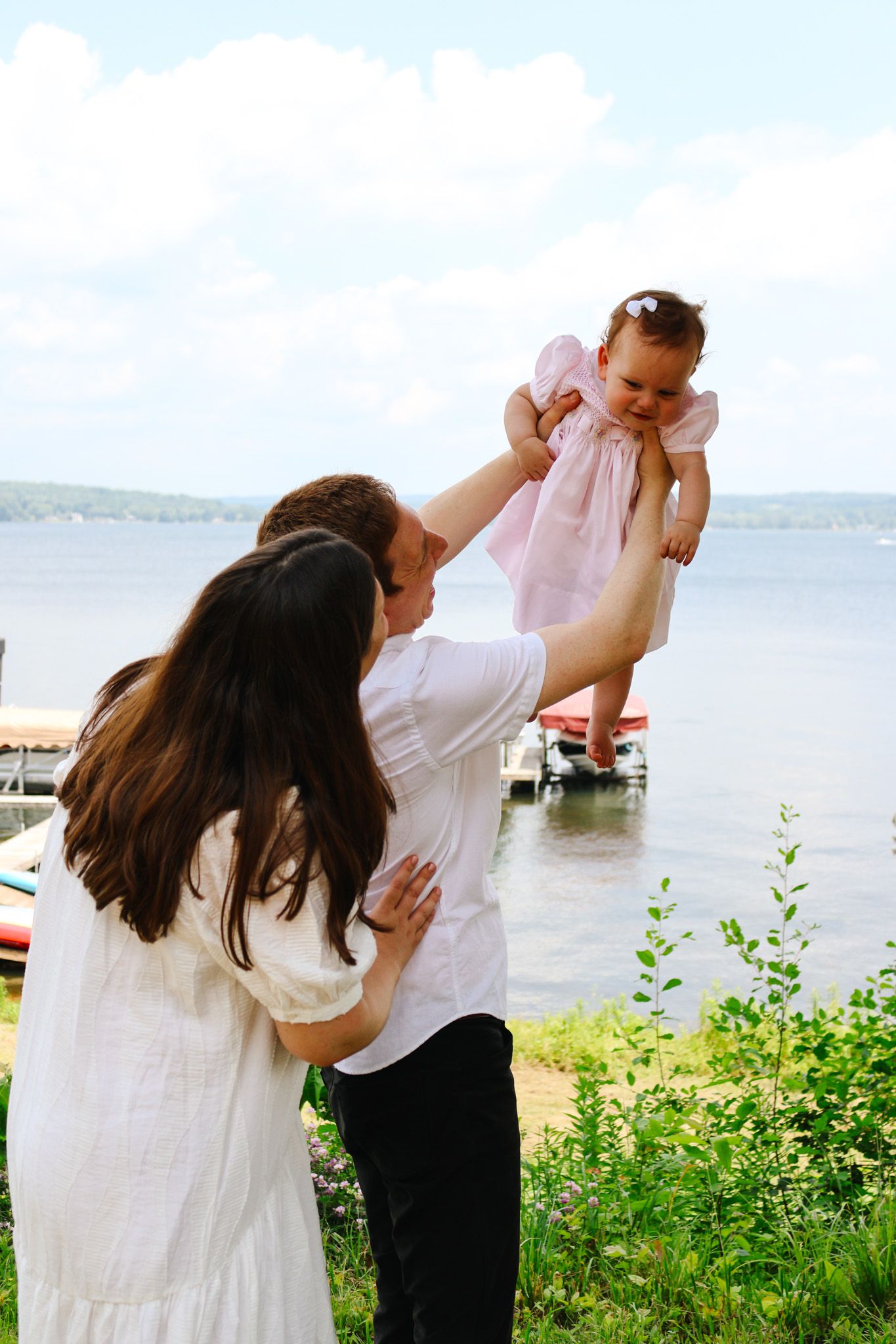 Parents joyfully lifting their laughing baby girl in a pink dress near a lakeside dock.