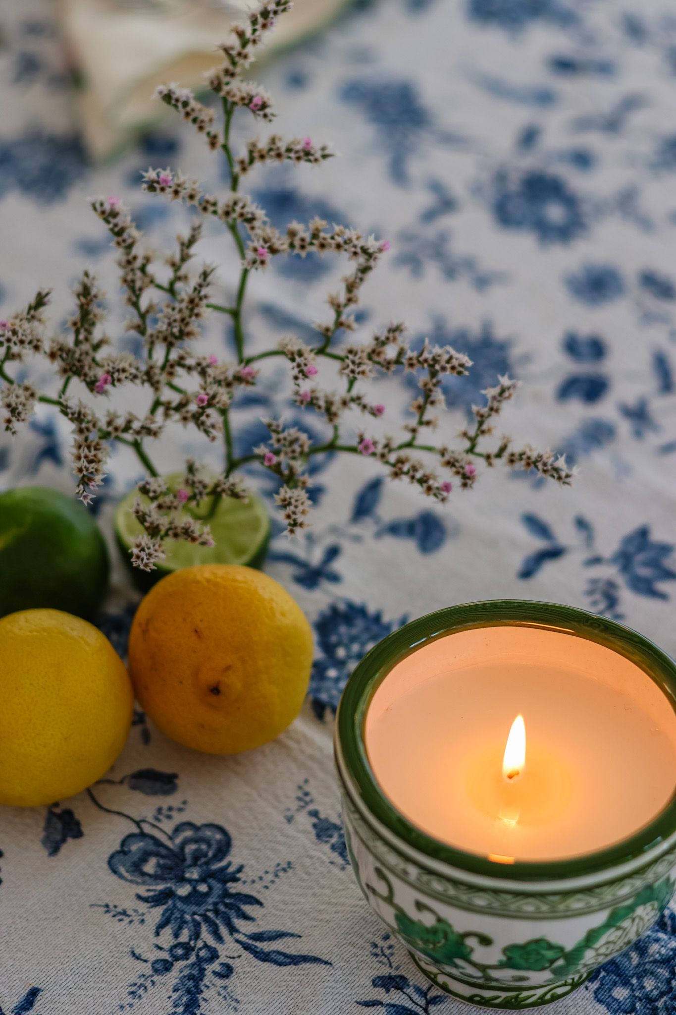 Lit green ceramic candle beside lemons and dried floral stems on blue floral tablecloth.