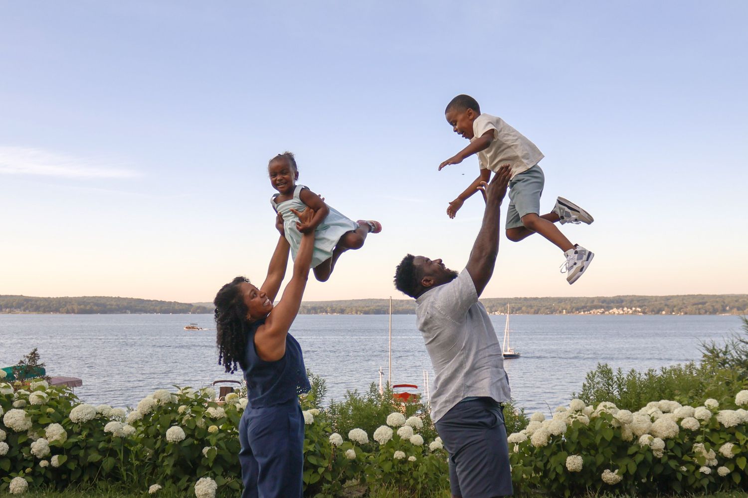 Joyful parents lifting two young children in the air near a waterfront garden at sunset.