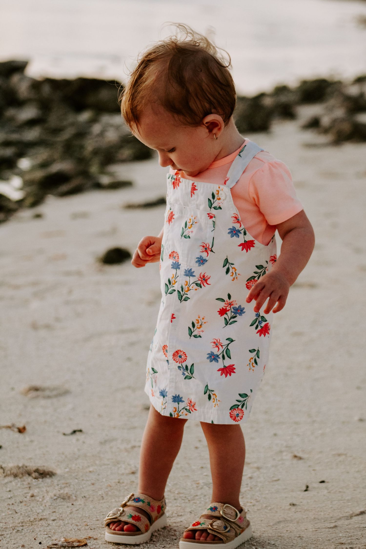Adorable toddler in a floral sundress and sandals standing on a sandy beach at sunset.