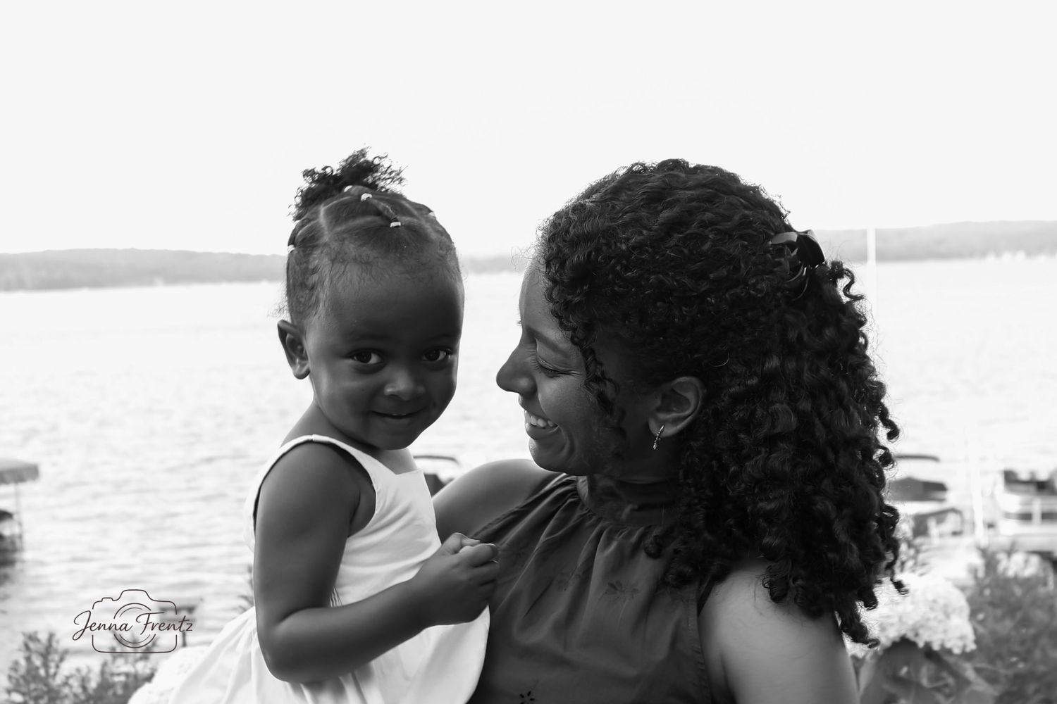 Black and white portrait of a mother tenderly holding her young daughter near a serene waterfront.