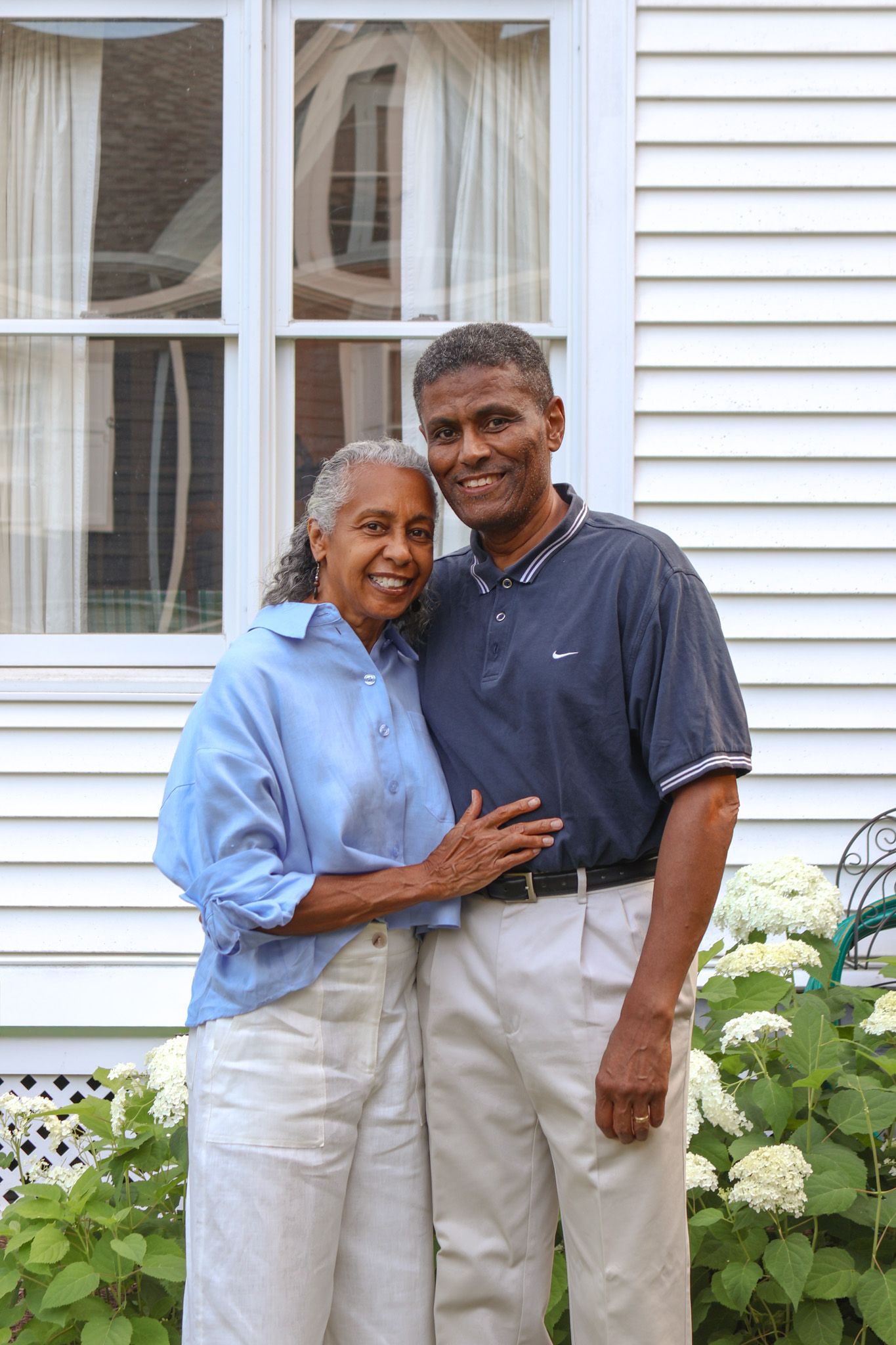Smiling senior couple embracing affectionately in front of a white house with hydrangea flowers.