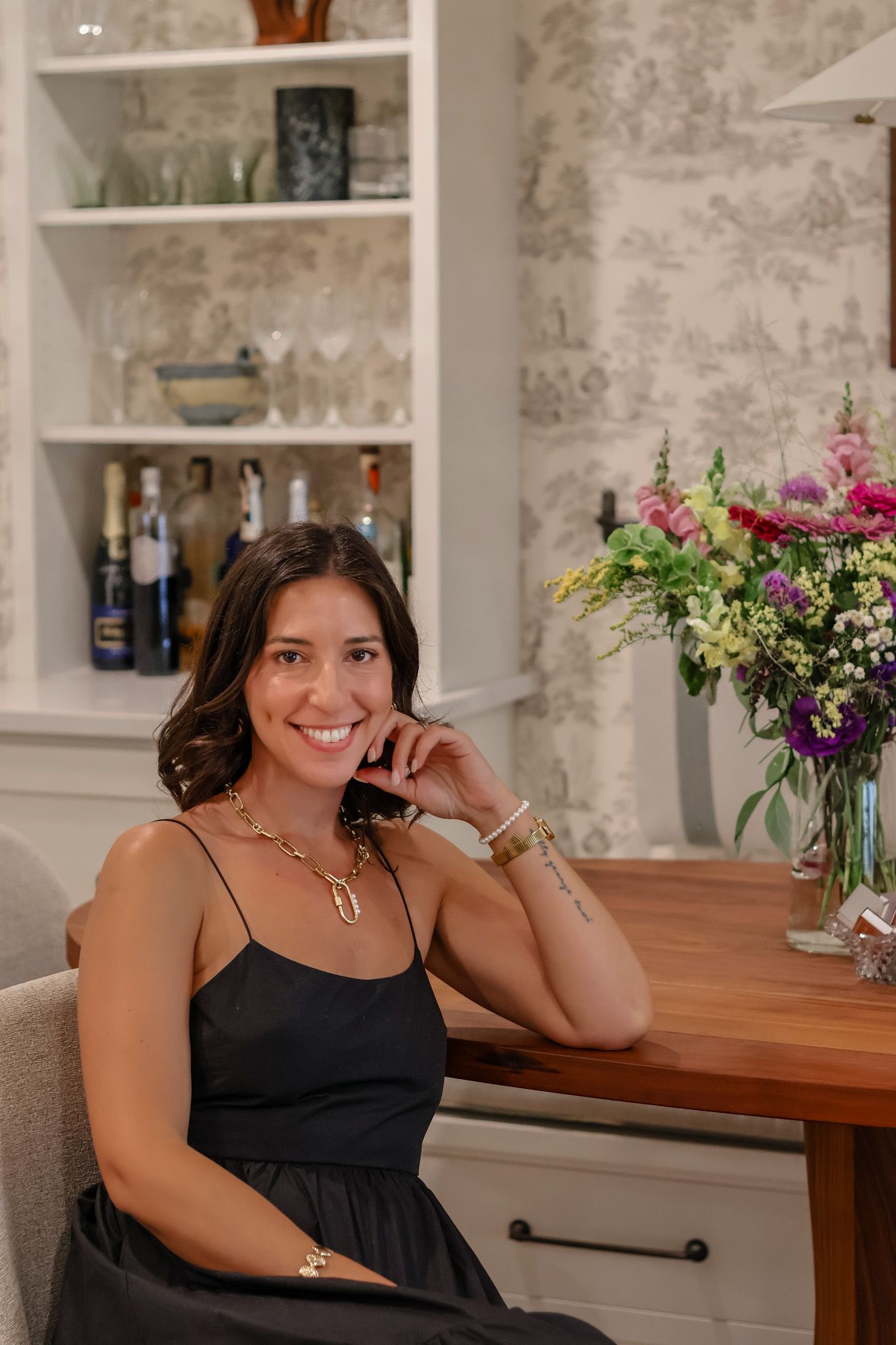 Smiling brunette woman in black dress seated indoors near floral arrangement and built-in shelving.