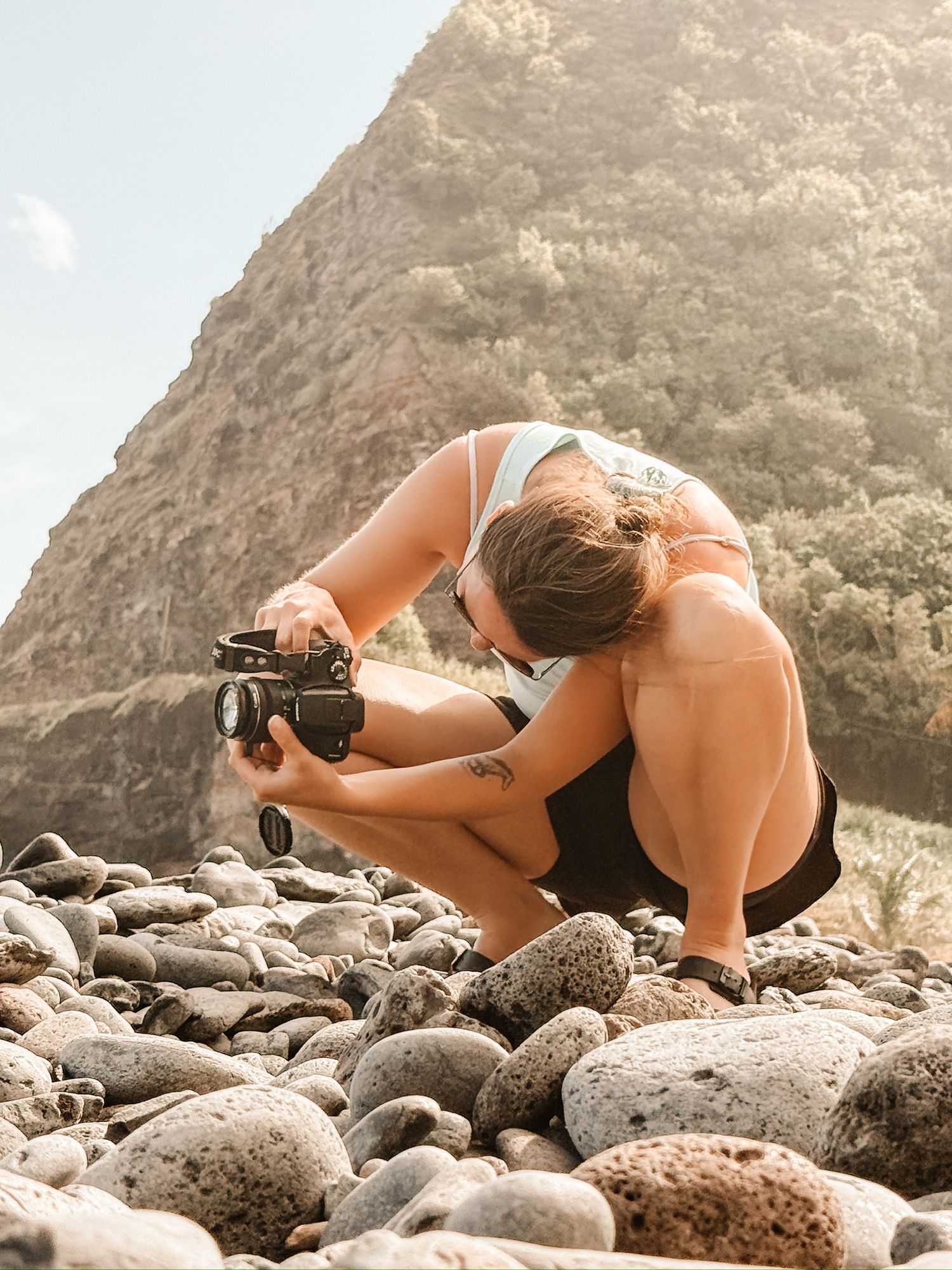 Woman crouching on rocky beach photographing with vintage camera near mountain backdrop.