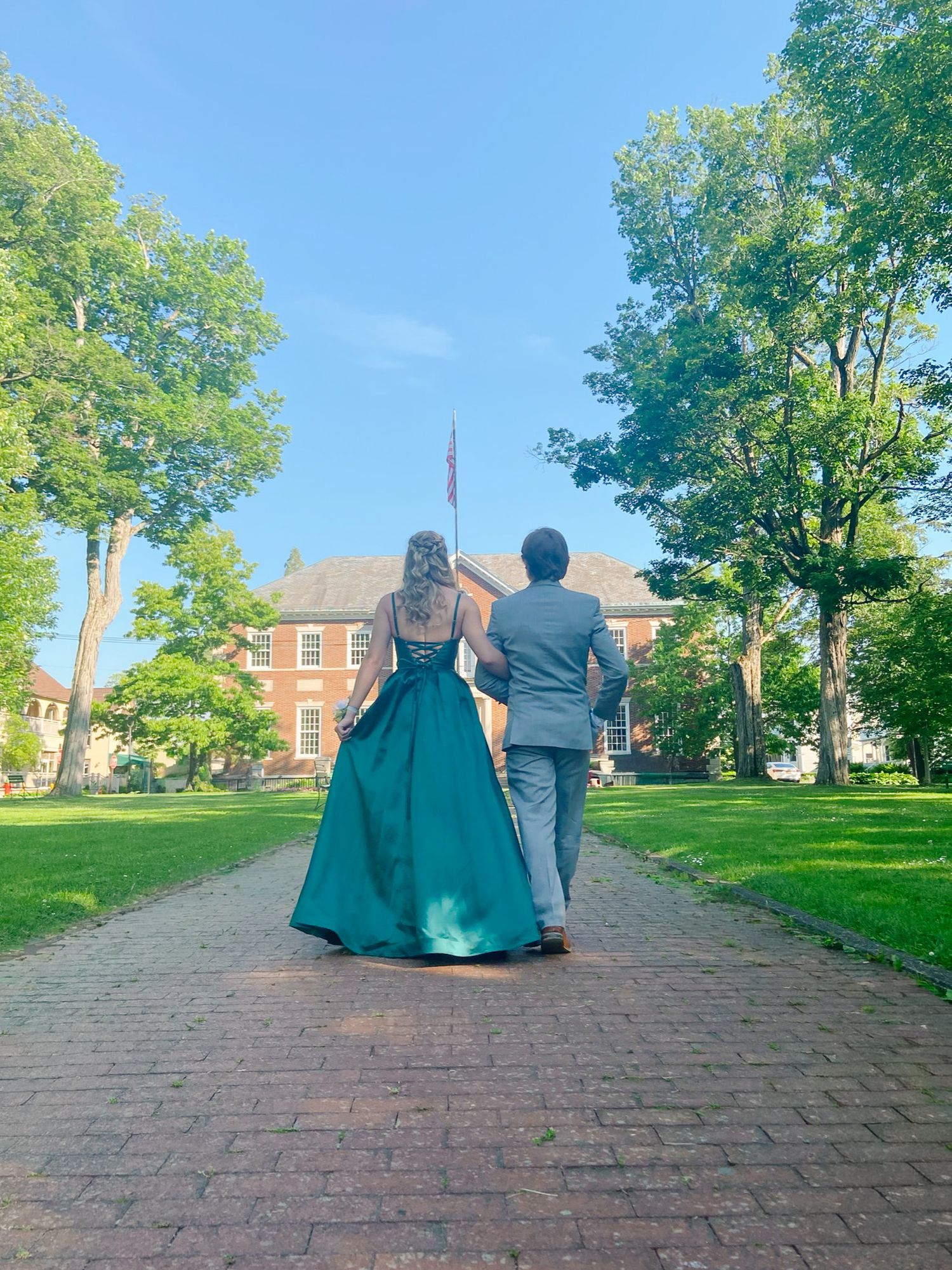 Couple in formal wear walking on brick path toward brick building surrounded by green trees on sunny day.