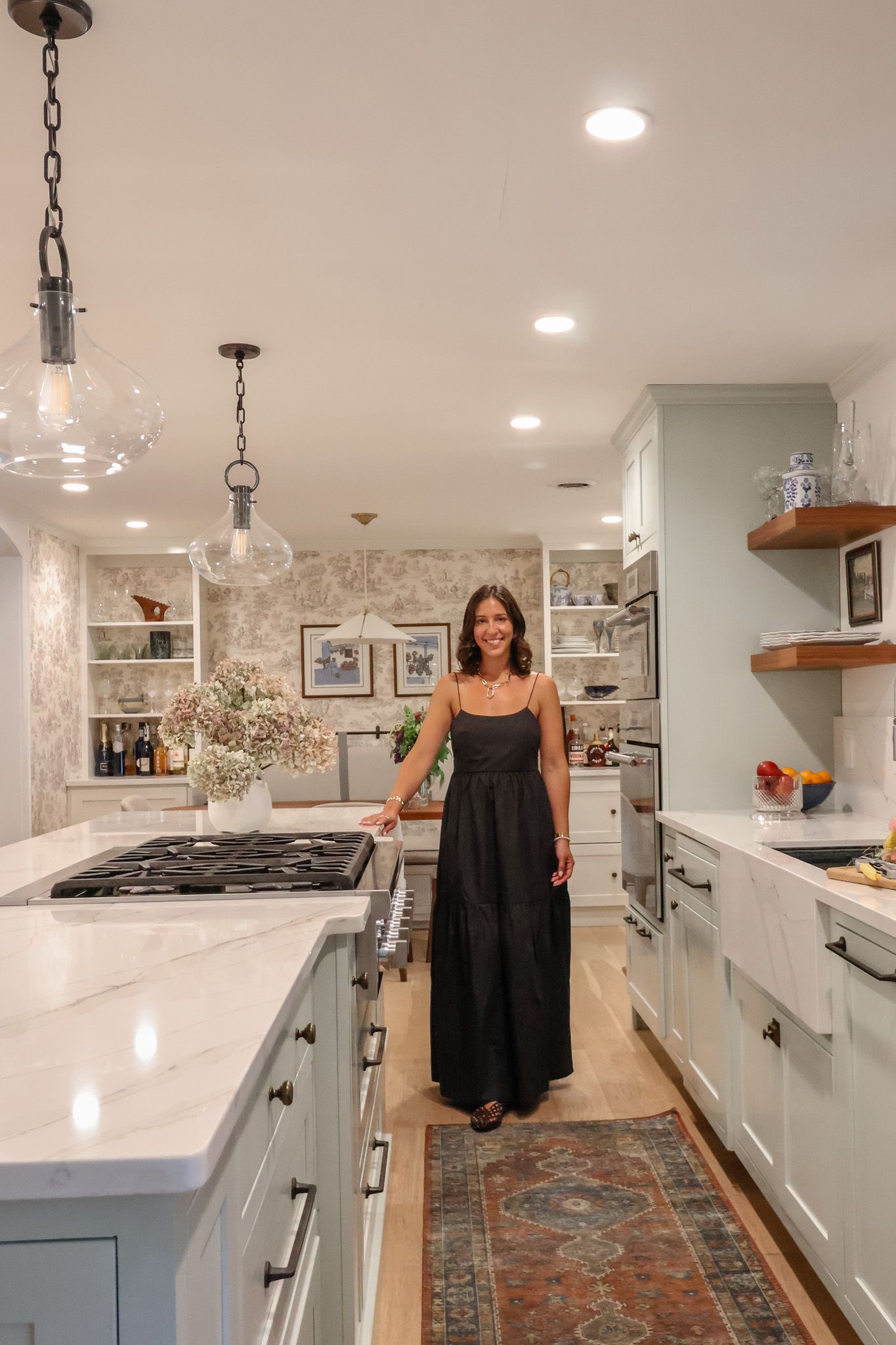 Smiling woman in black maxi dress standing in elegant renovated kitchen with sage green island.