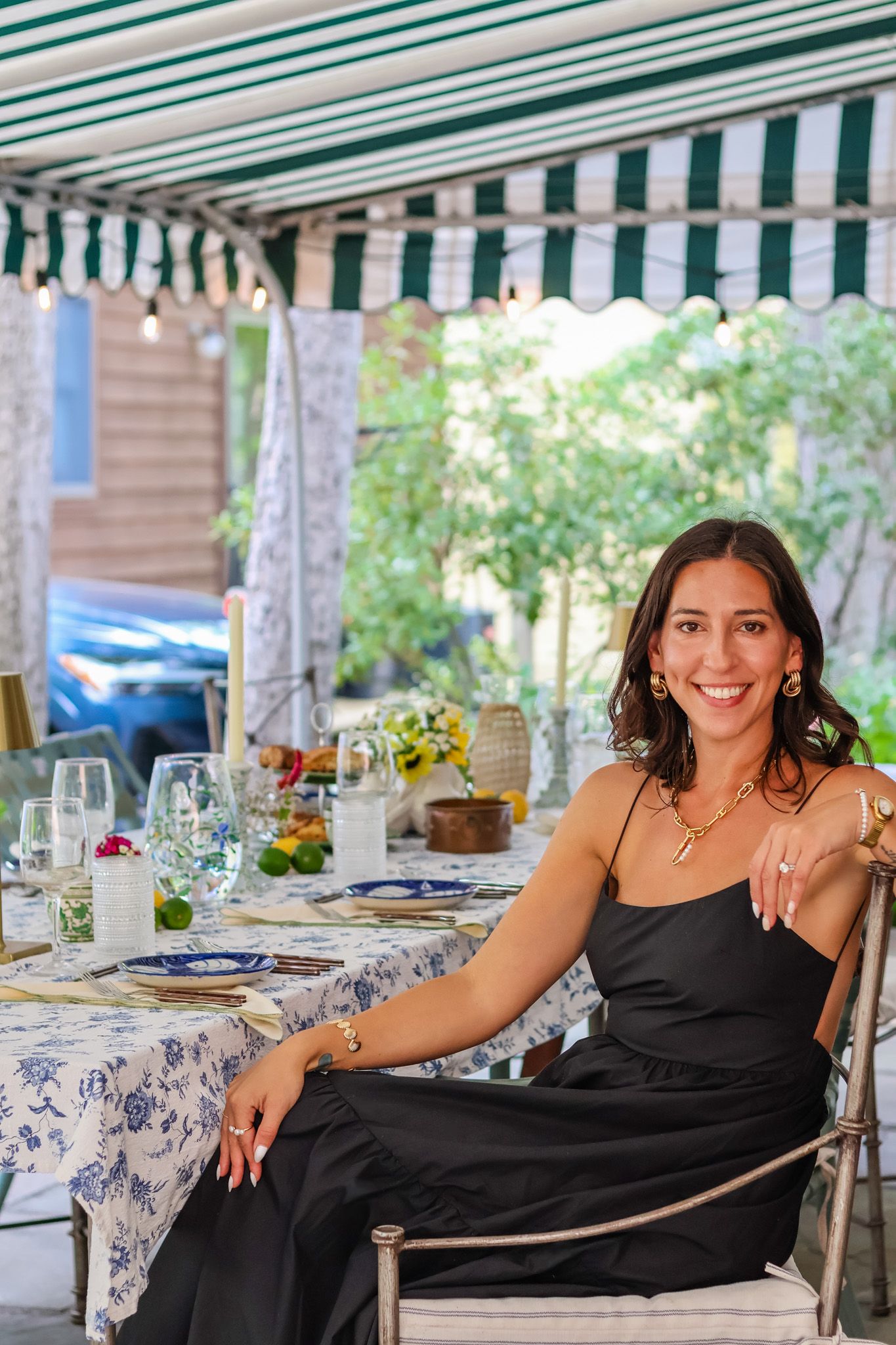 Smiling woman in a black dress seated at an elegantly set outdoor dining table under a striped awning.
