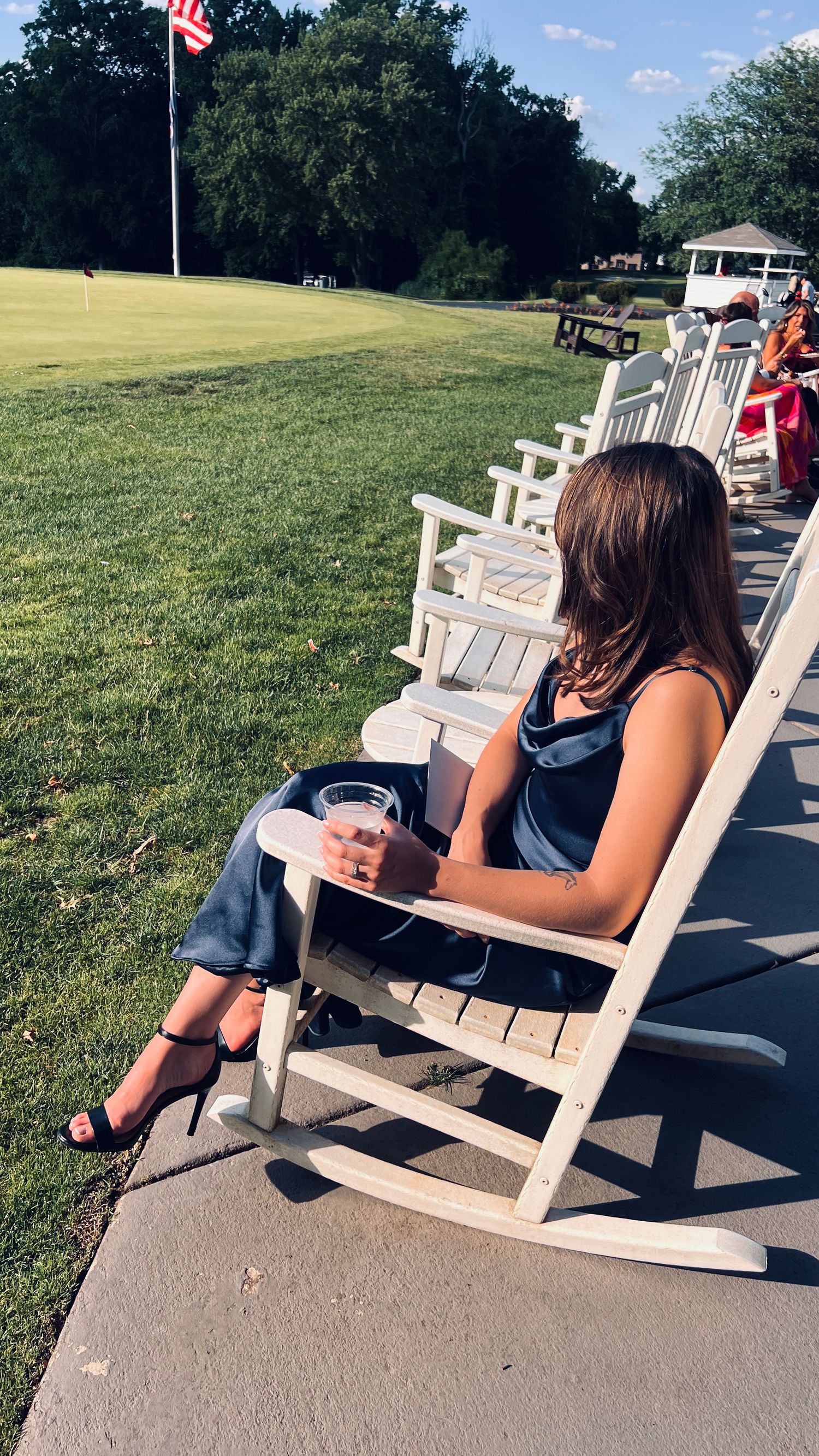 Woman in a navy dress relaxing in a white rocking chair on a sunny green lawn holding a drink.