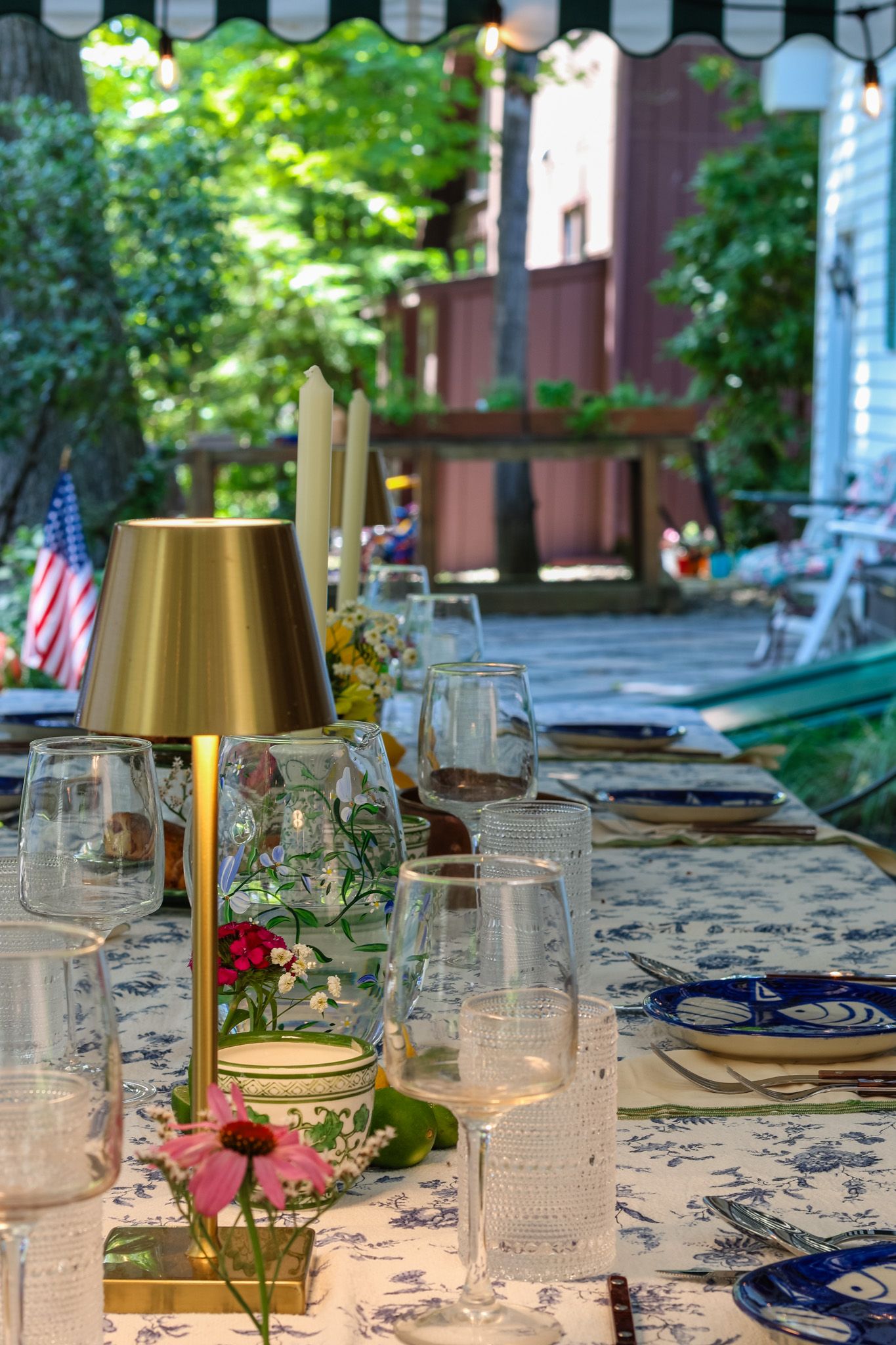 Elegant outdoor dining table set with glassware, gold lamp, and blue floral tablecloth in garden.