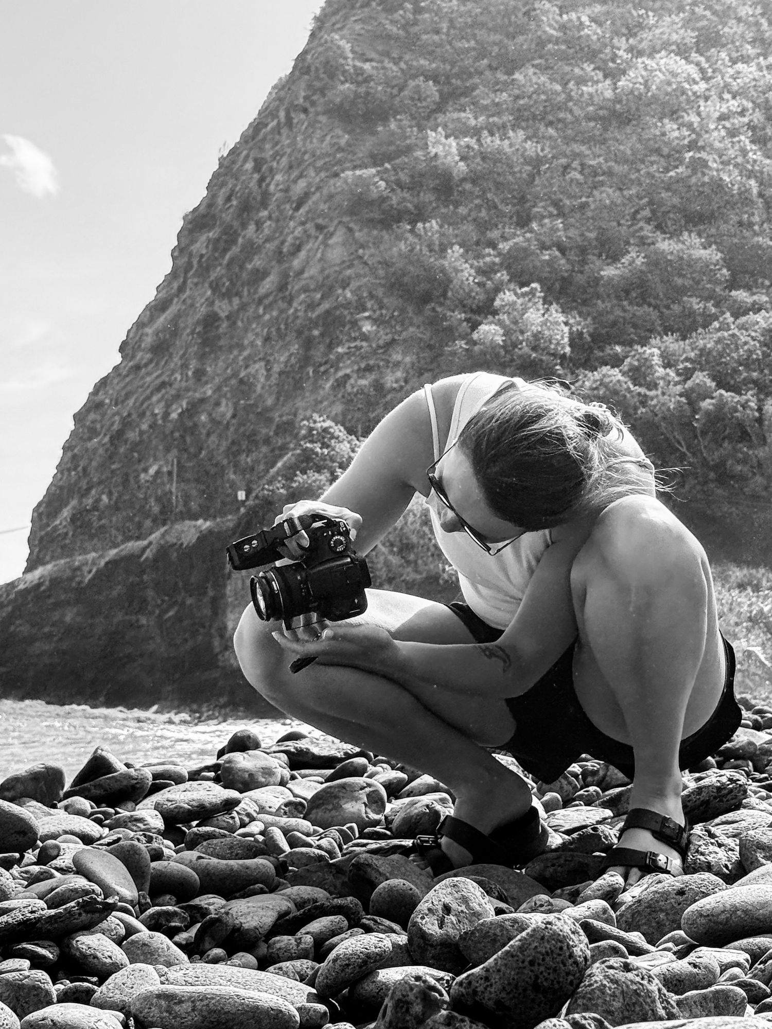 Black and white photo of a woman crouching on a rocky beach photographing with a camera near a cliff.