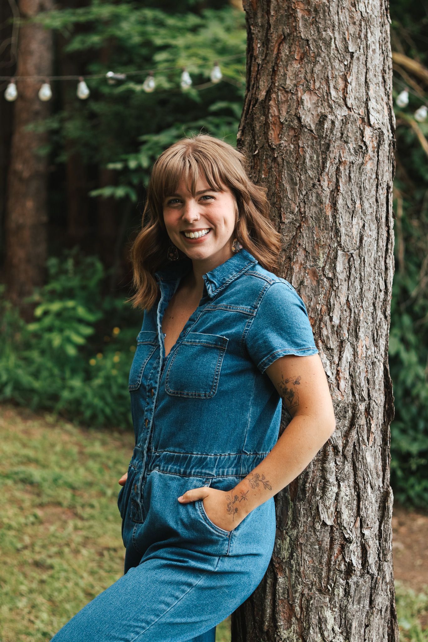 Smiling woman in denim jumpsuit leaning against a tree in a lush green outdoor setting.