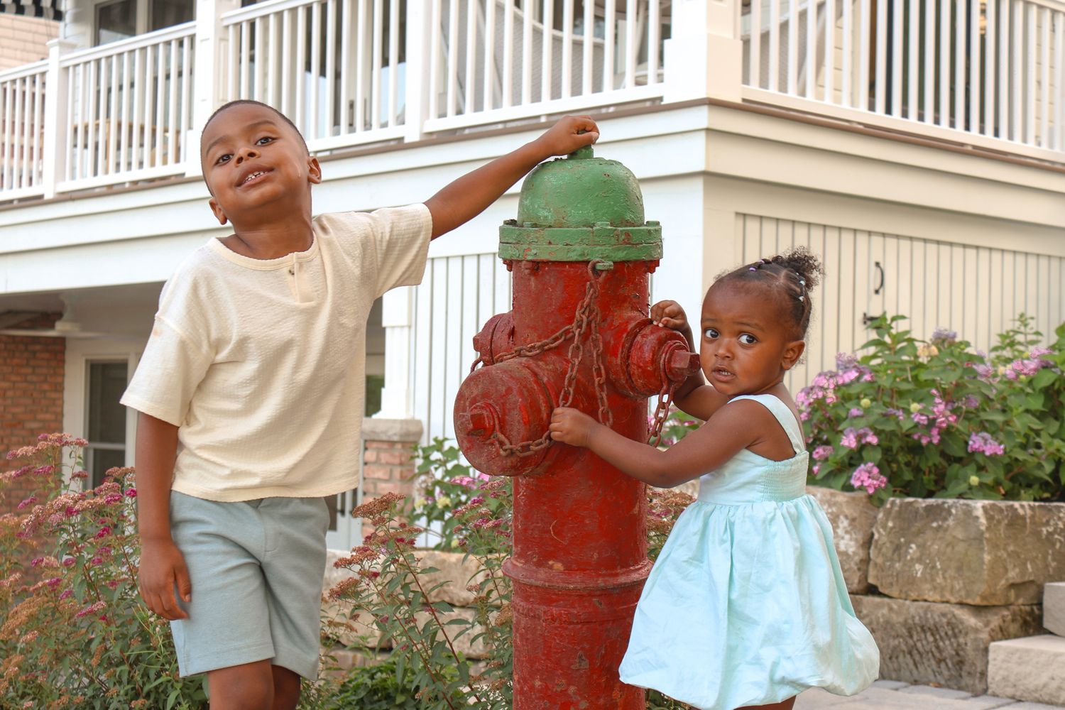 Two young children smiling beside a red fire hydrant outside a residential building with pink flowers.