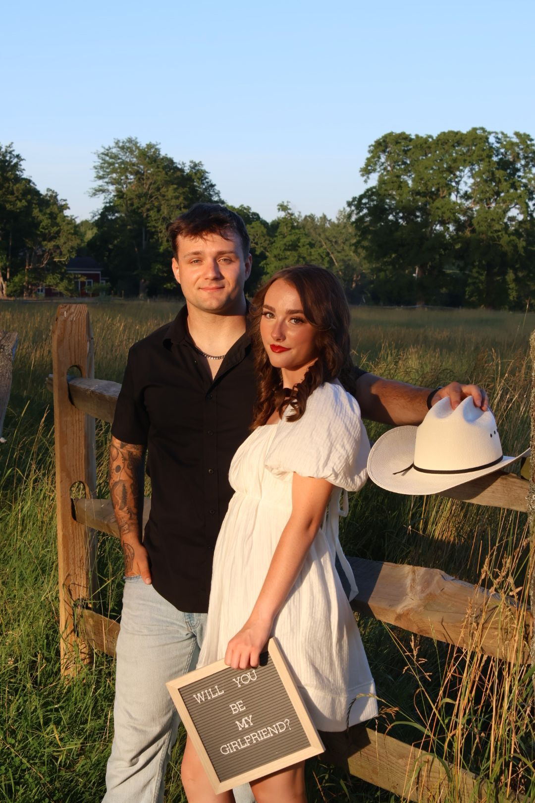 Couple posing at wooden fence holding Will You Be My Girlfriend sign at golden hour.