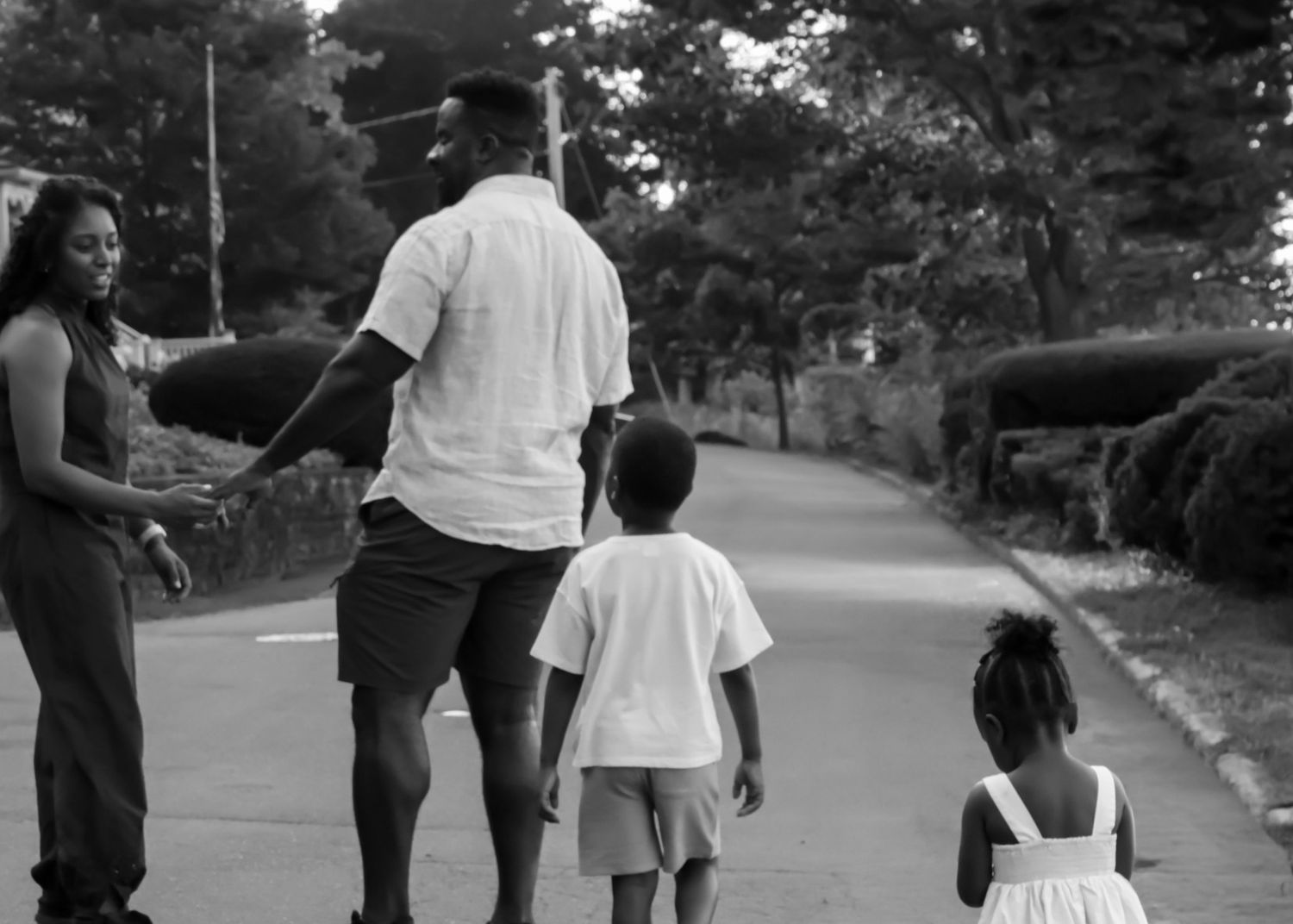 Black and white photo of a family walking together down a tree-lined road, parents holding hands with young children.