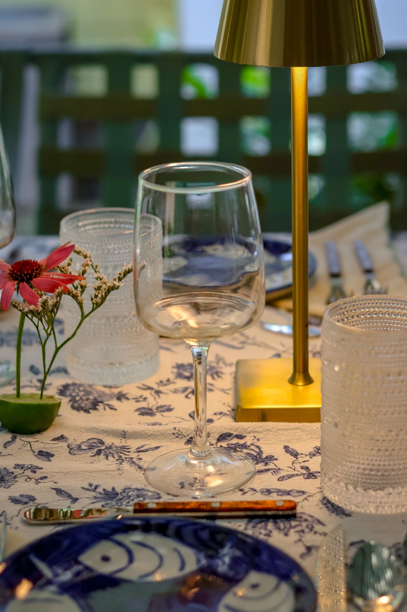 Crystal wine glass and gold table lamp on blue floral tablecloth at elegant outdoor dinner setting.