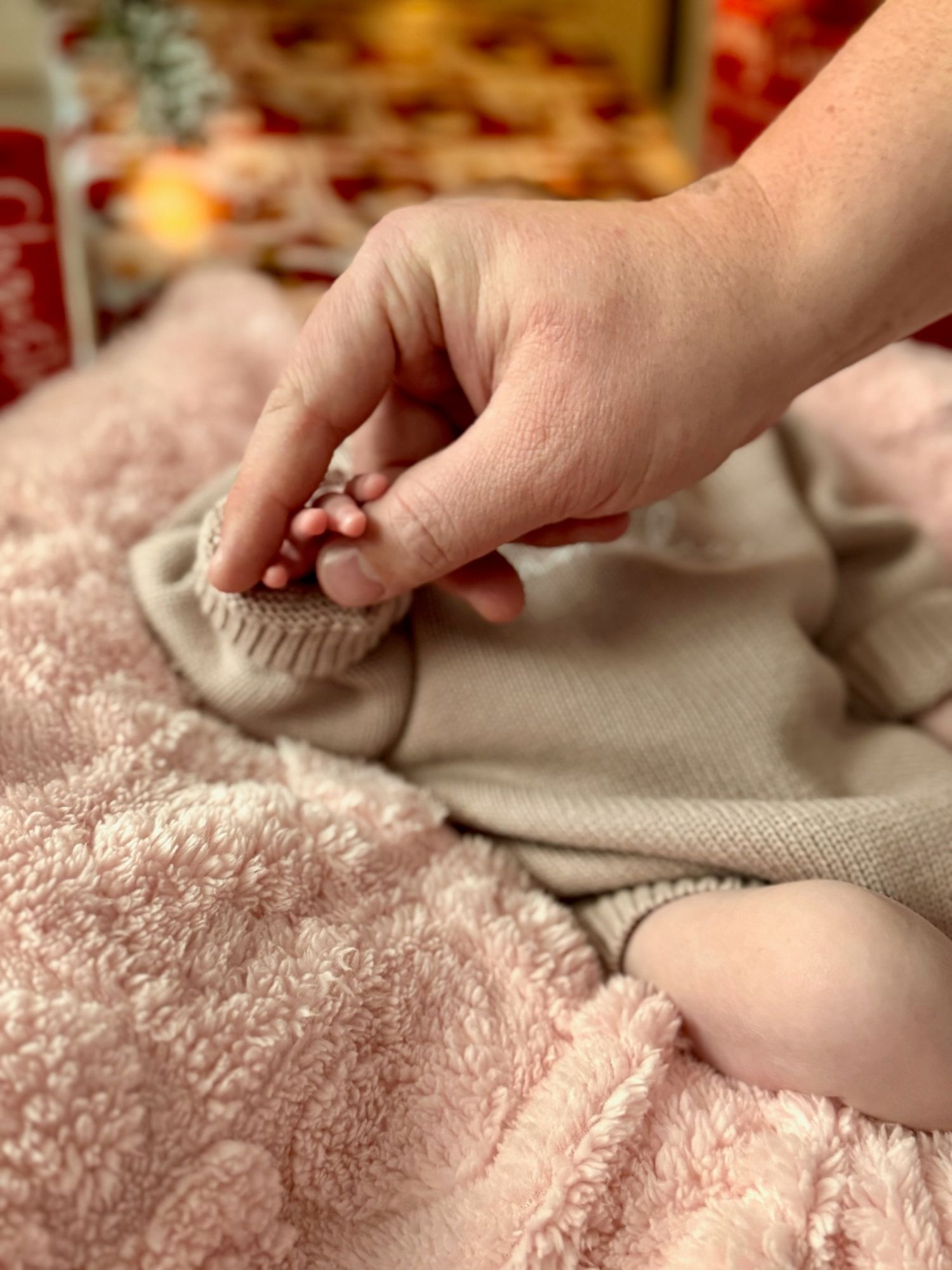Adult hand gently holding tiny baby toes on a soft pink blanket near Christmas decor.