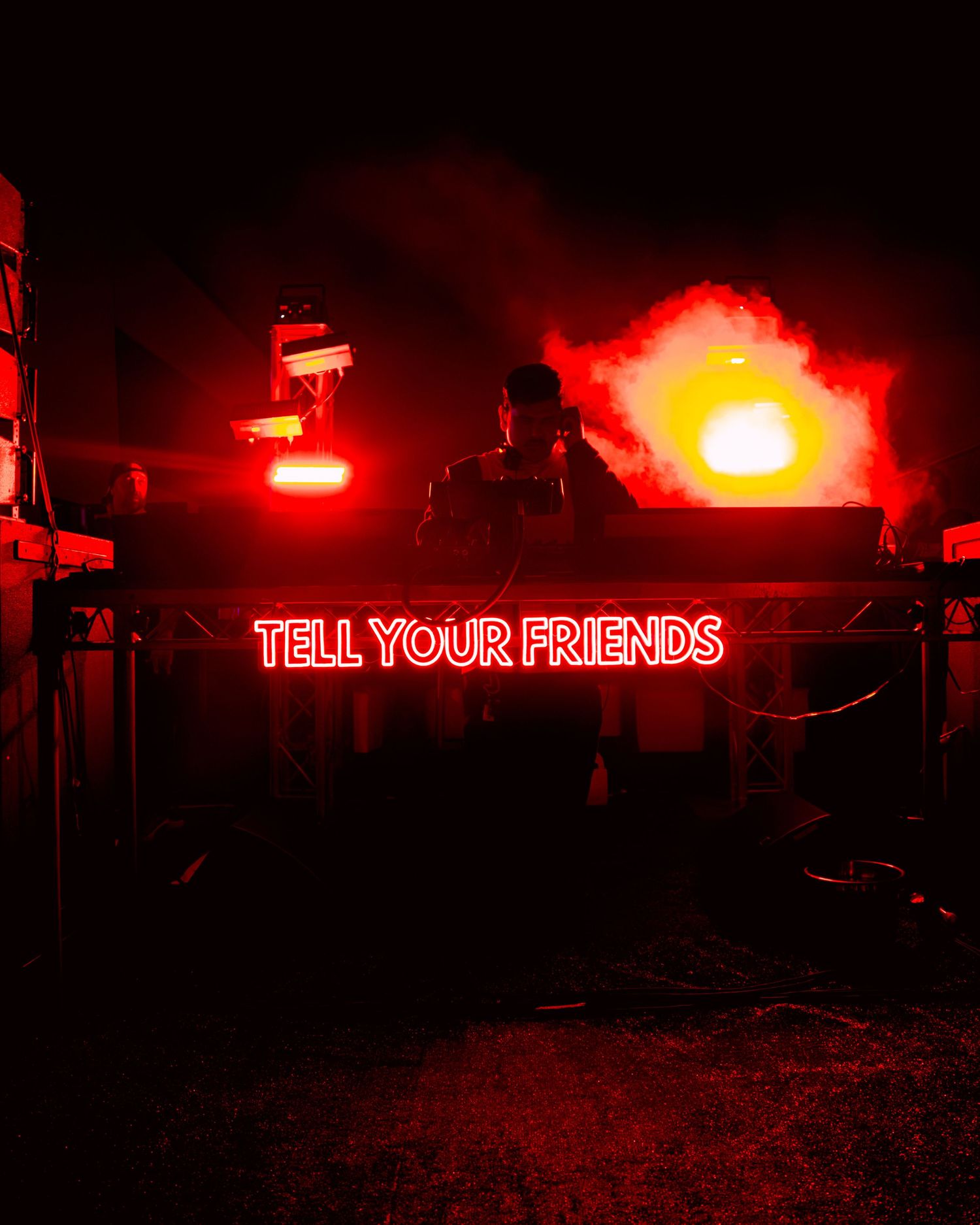 DJ performs behind a neon sign reading Tell Your Friends with red smoke and lights.