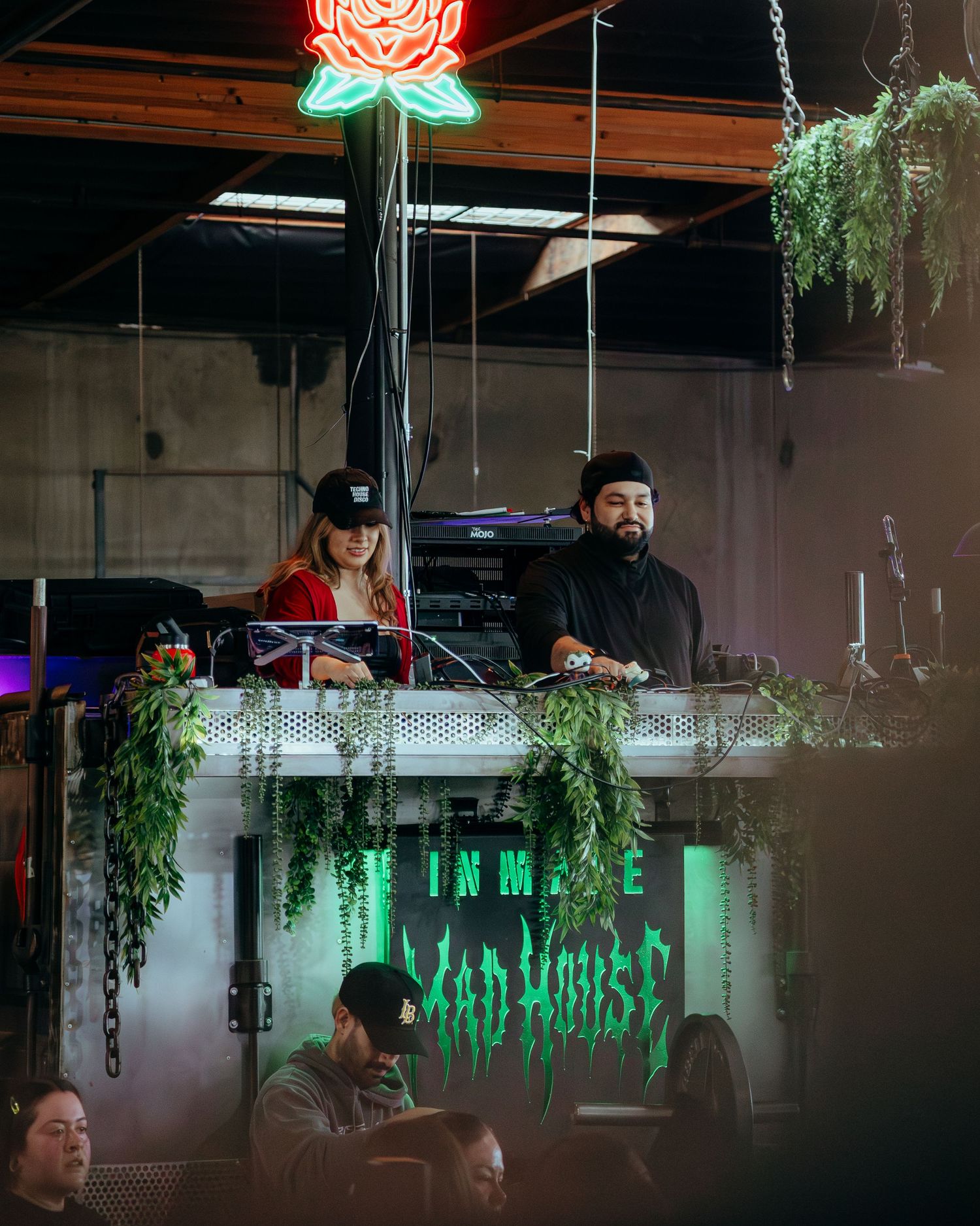 Two DJs perform at a venue with neon signs and hanging plants under a glowing rose neon light.