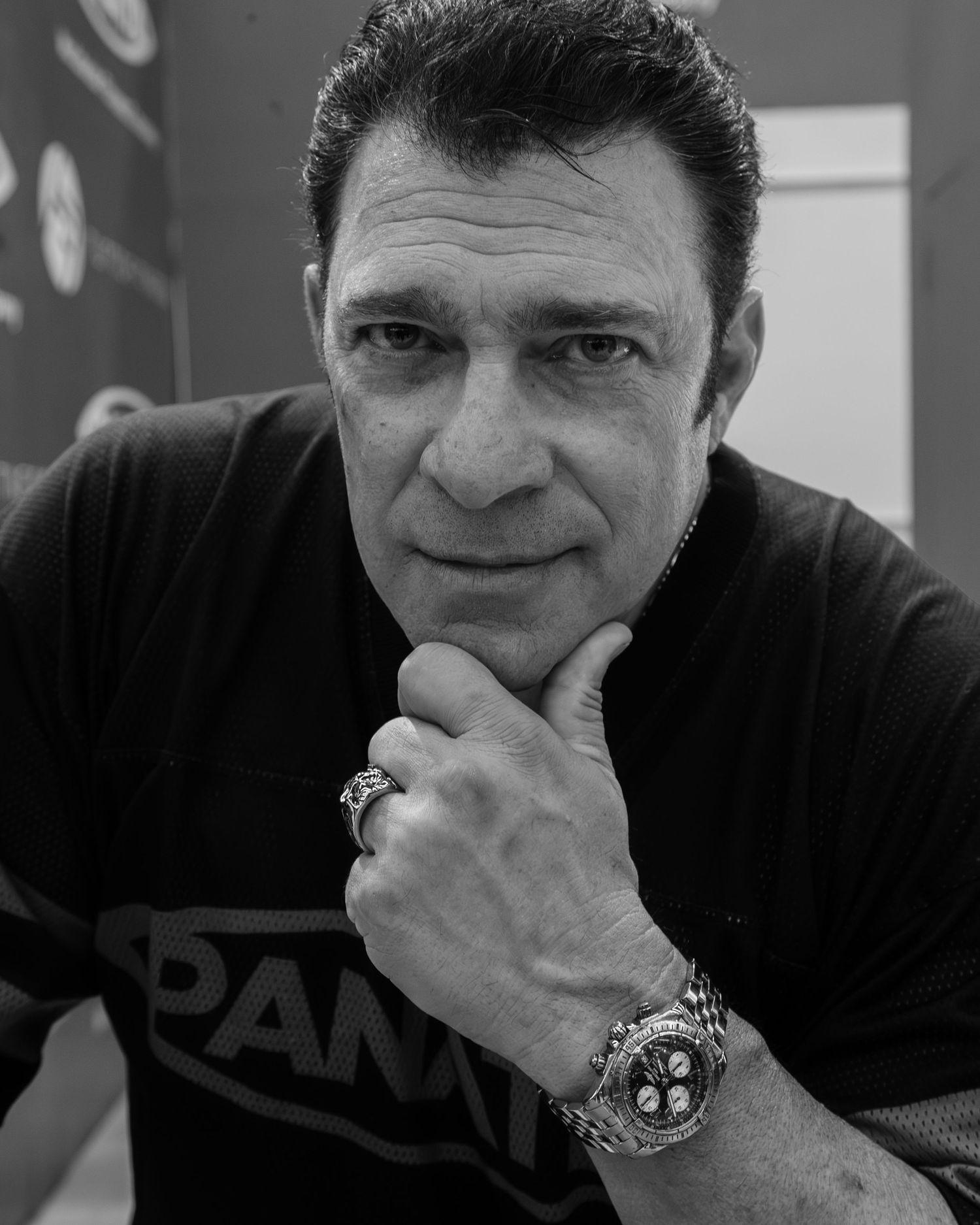 Black and white portrait of a middle-aged man in a SPAM t-shirt, resting chin on hand, wearing a watch and ring.