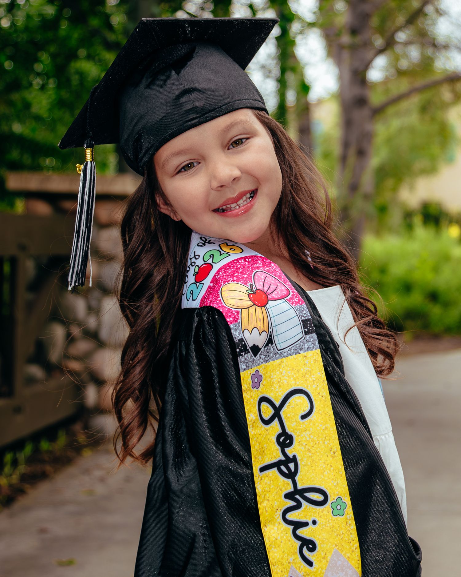Young girl smiling in black graduation cap and gown with personalized yellow sash reading Sophie outdoors.
