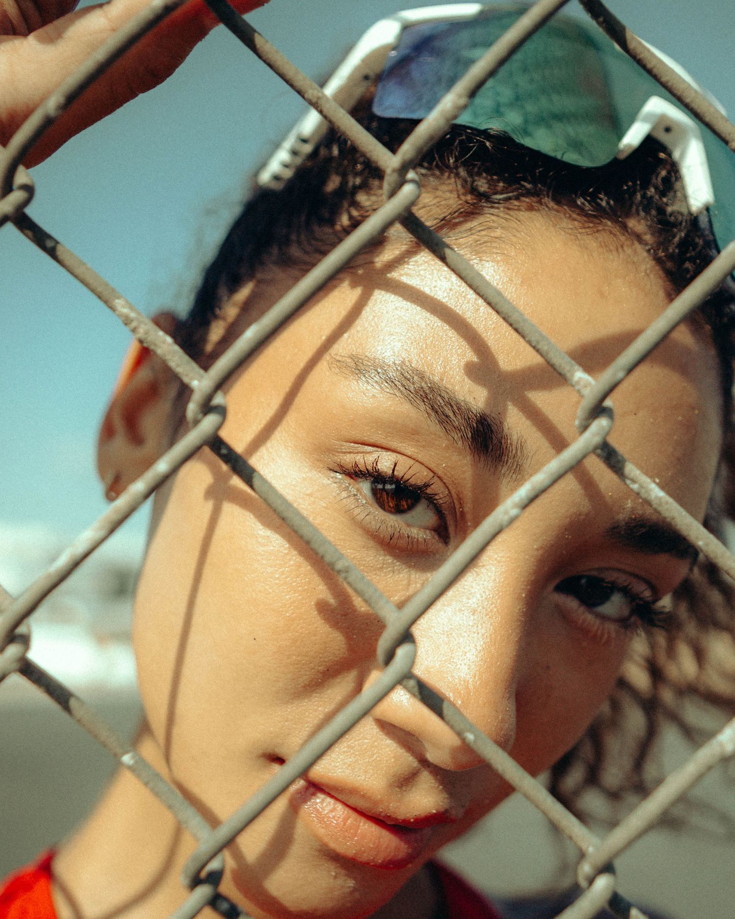 Close-up portrait of young woman with curly hair behind chain-link fence in warm sunlight.