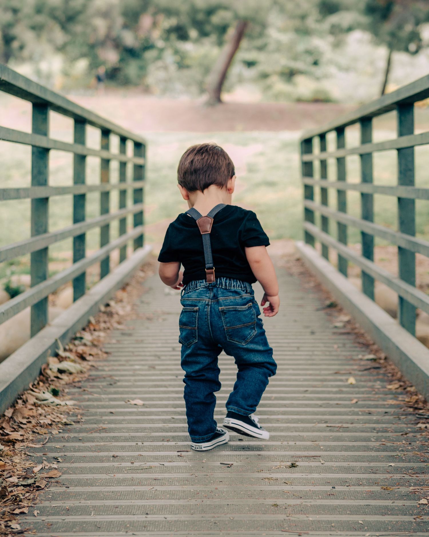 Toddler boy in suspenders and jeans walks across wooden bridge in a lush green park.