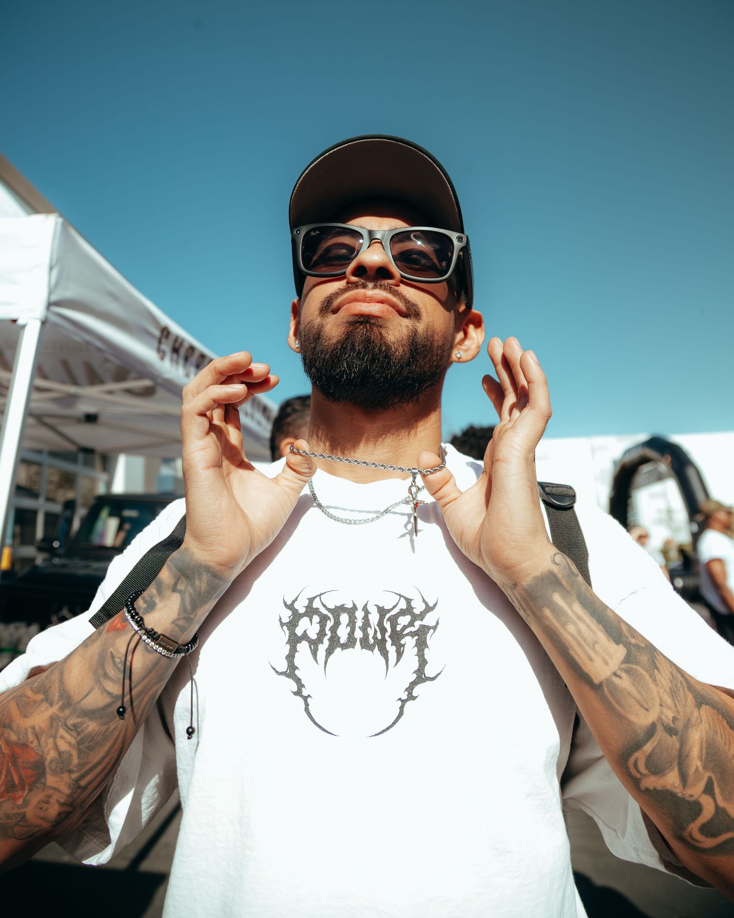 Tattooed man in white graphic tee and cap adjusts necklace outdoors on a sunny day.