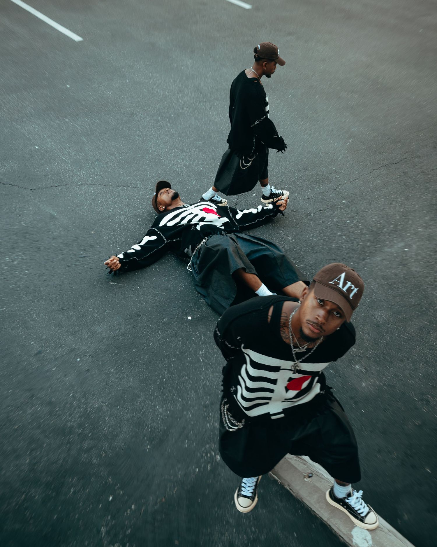 Three young men in skeleton print streetwear posing dramatically on a parking lot from aerial view.