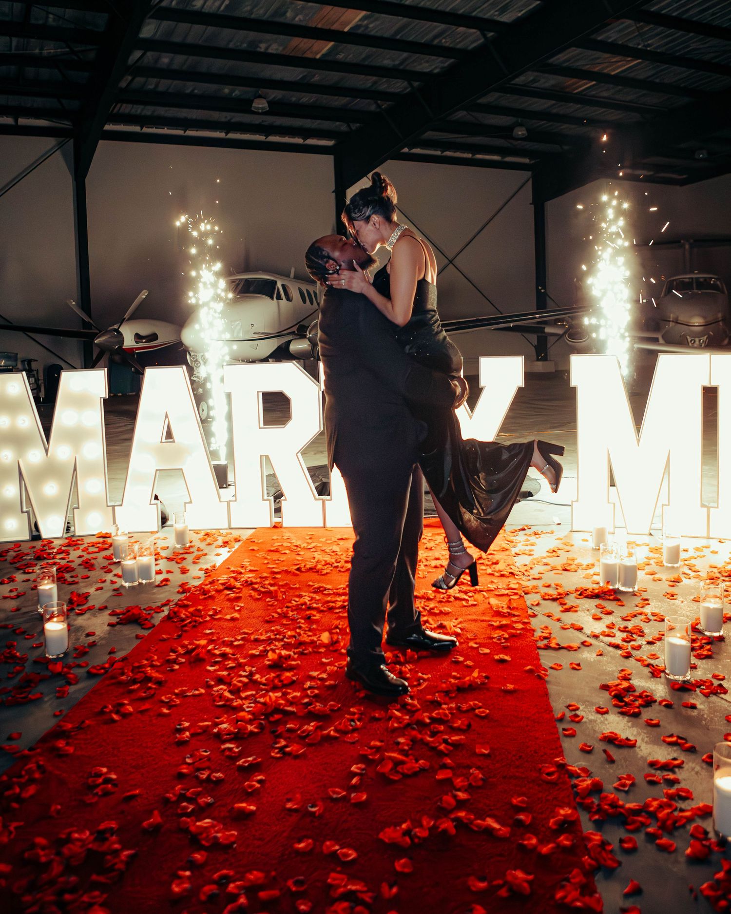 Couple sharing a kiss at a stunning marry me proposal setup with sparklers in a hangar.