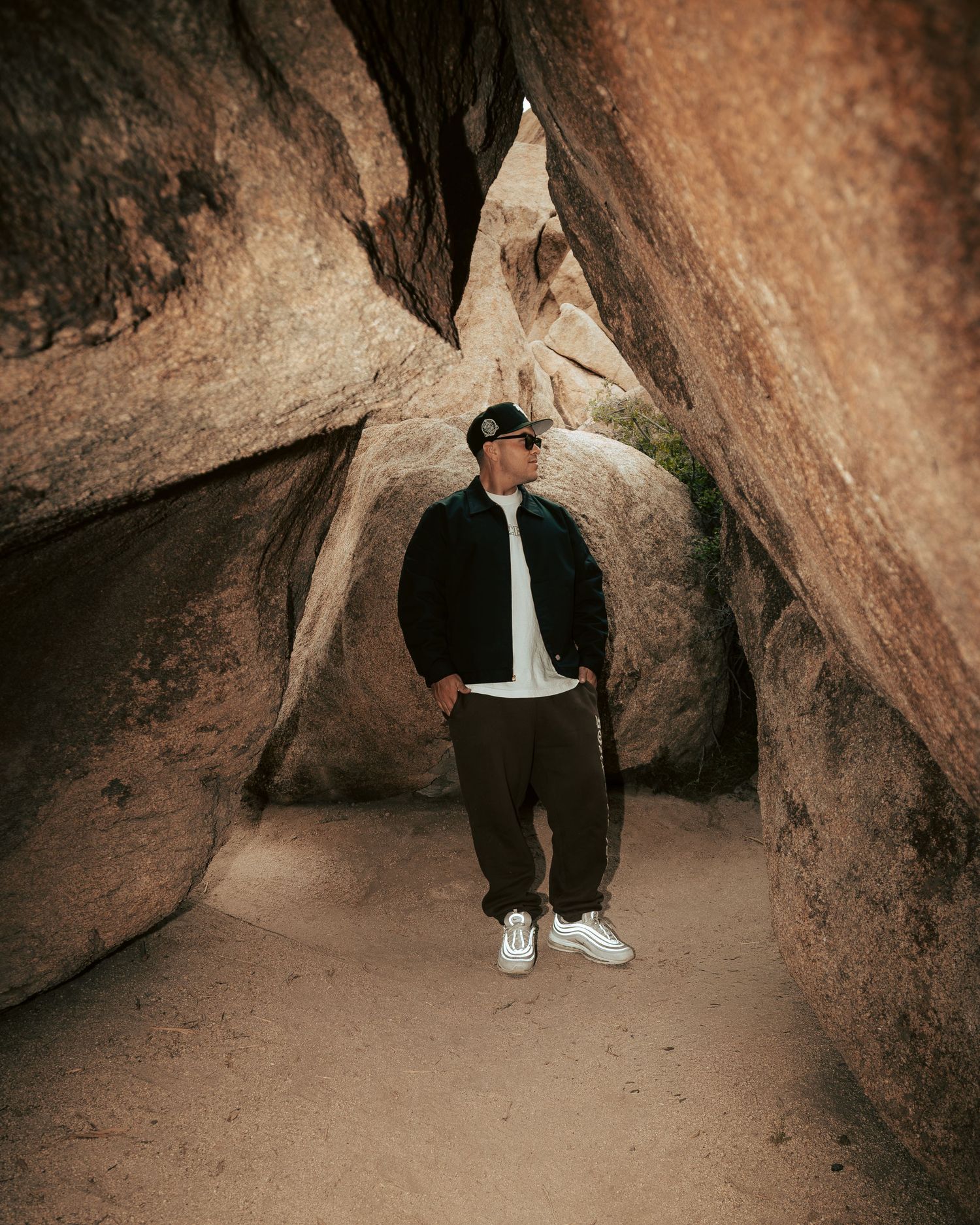 Man in dark jacket and cap standing between large boulders on sandy desert trail.