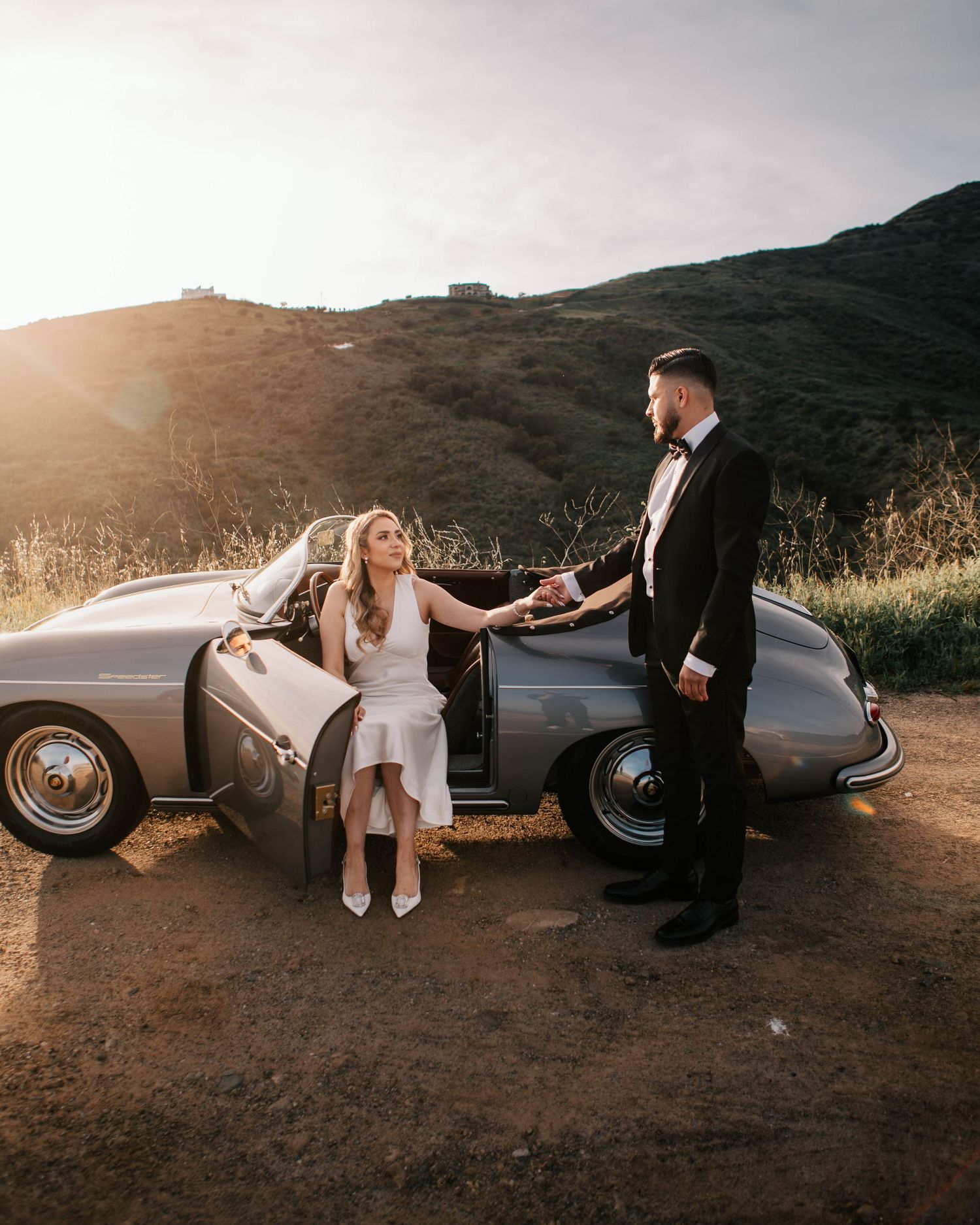 Elegant couple in wedding attire poses with vintage silver sports car at golden sunset hillside.