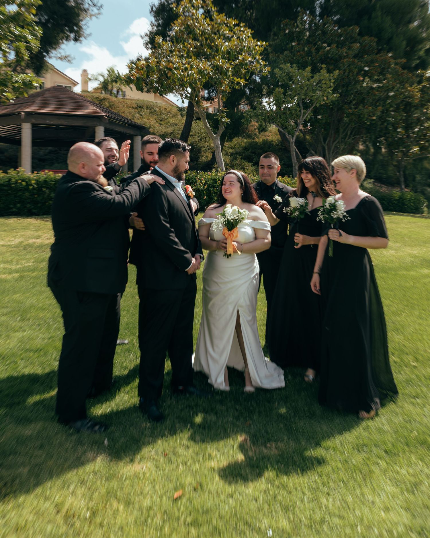 Wedding party in black formal attire surrounds smiling bride and groom on lush green lawn outdoors.