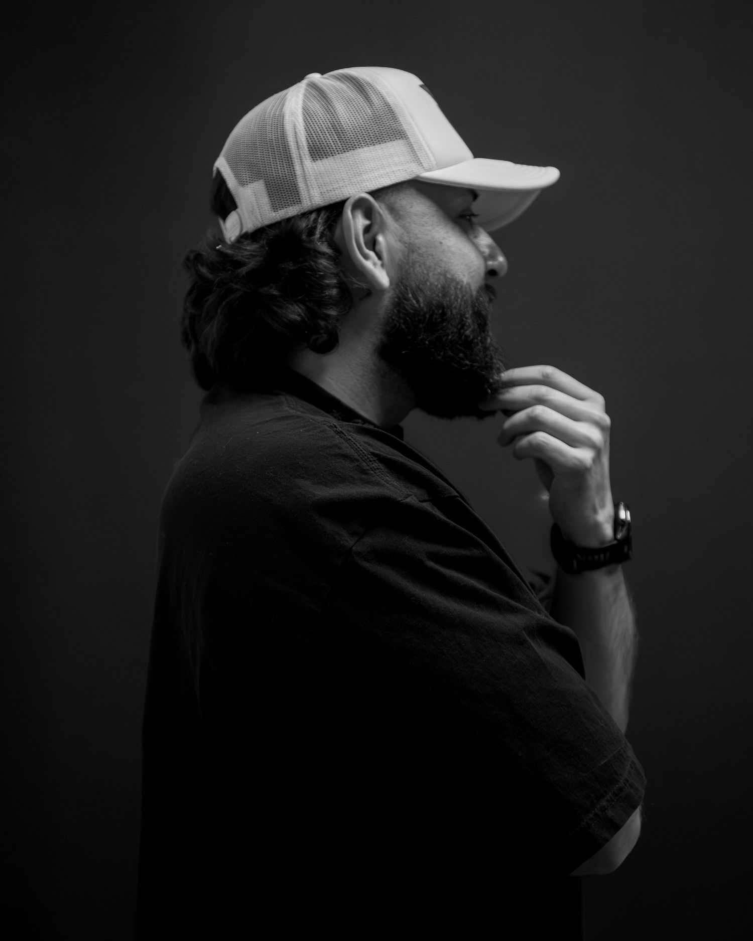 Black and white side profile portrait of bearded man in white trucker cap on dark background.