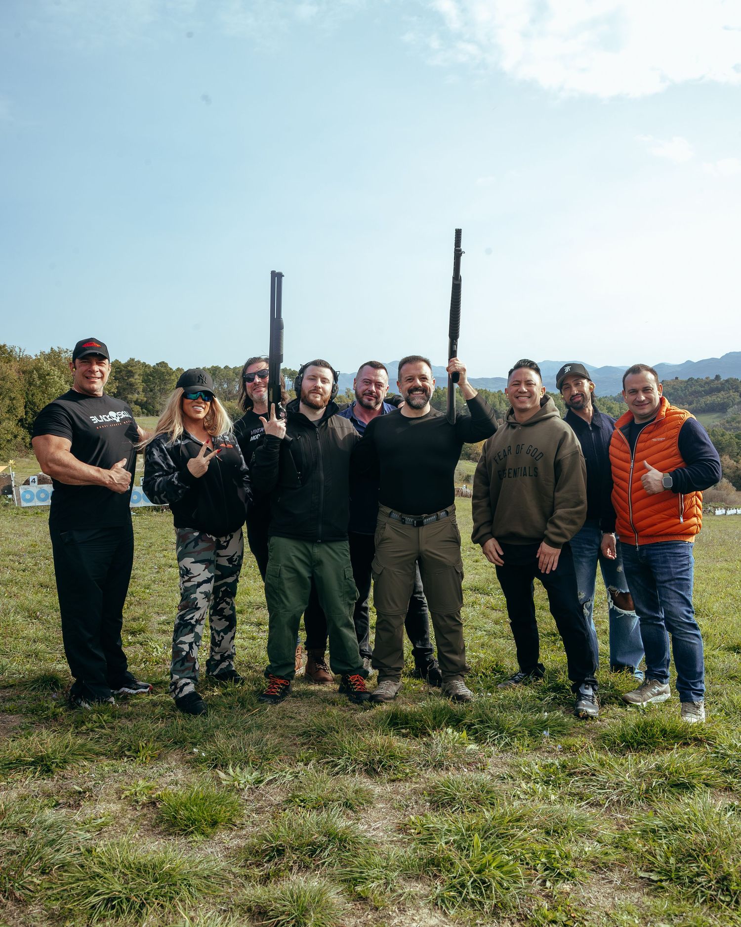 Group of nine people pose outdoors holding shotguns at a clay shooting range.