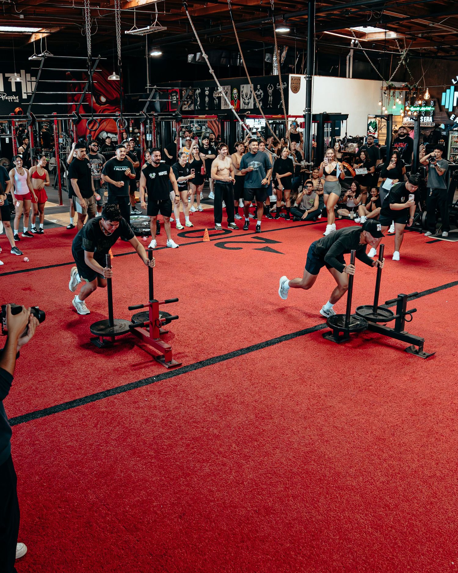 Two athletes push weighted sleds on red turf floor at an indoor gym competition with crowd watching.