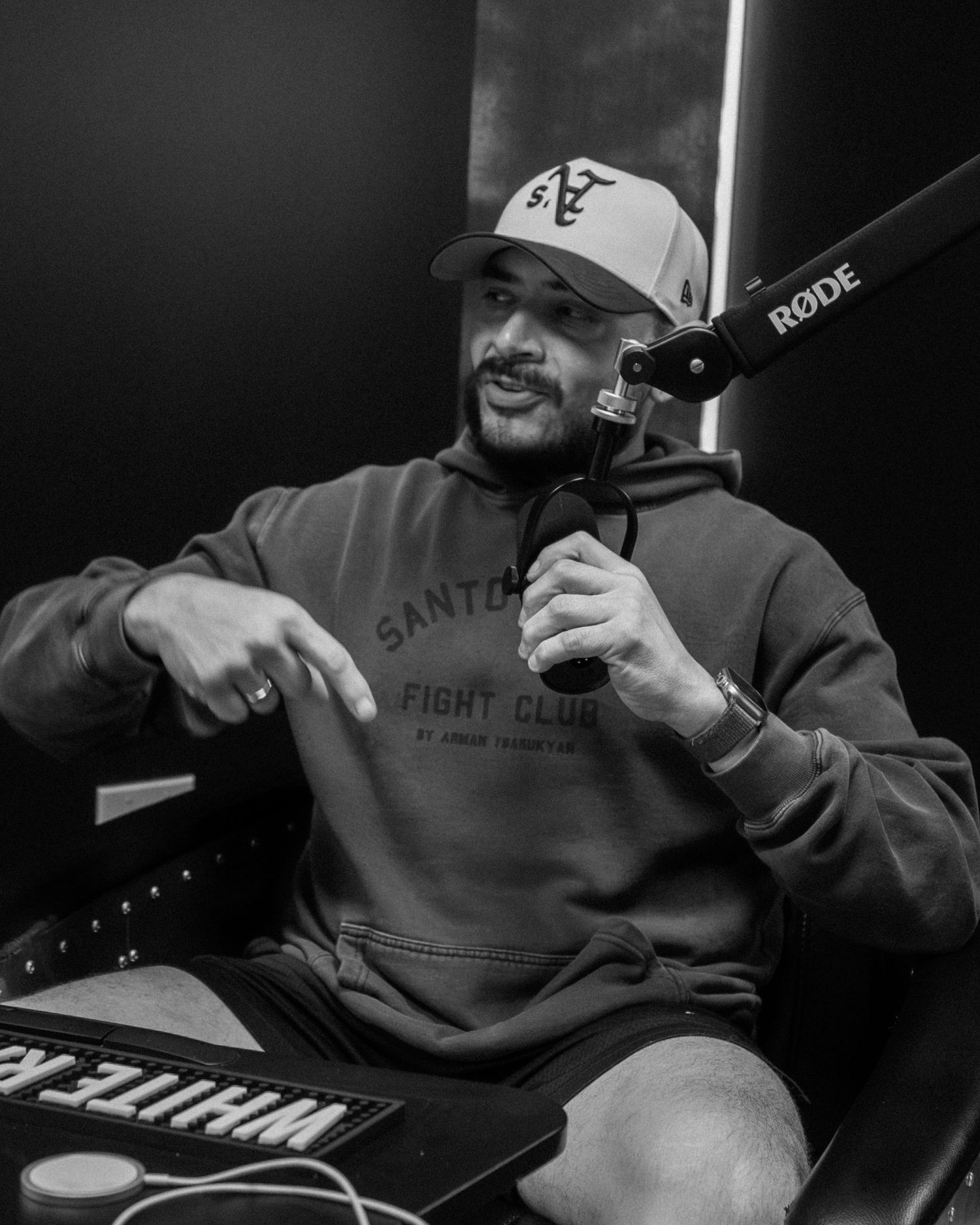 Black and white photo of a man in a hoodie and cap speaking into a Rode microphone in a studio.