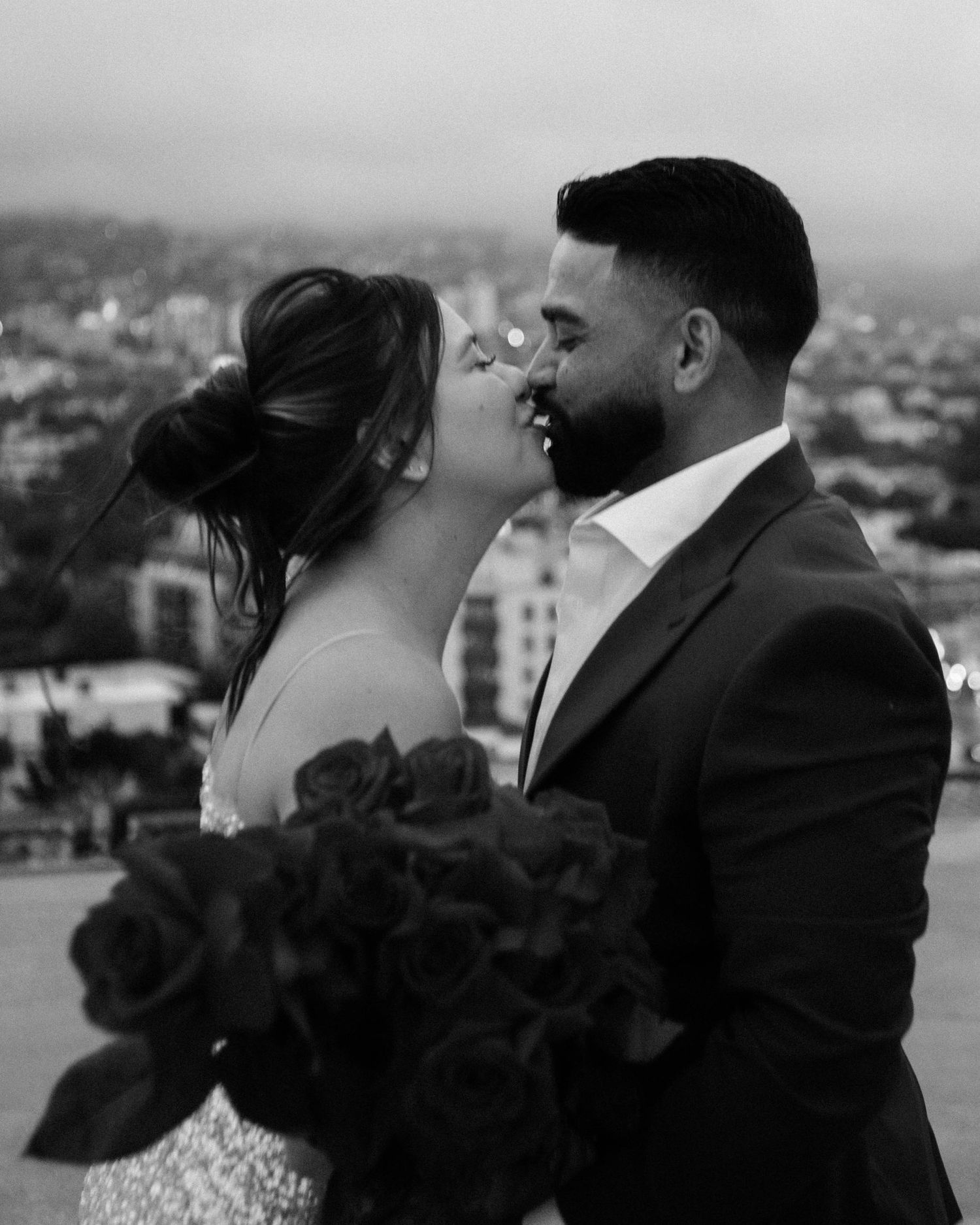 Black and white photo of a couple almost kissing with a bouquet of dark roses outdoors.