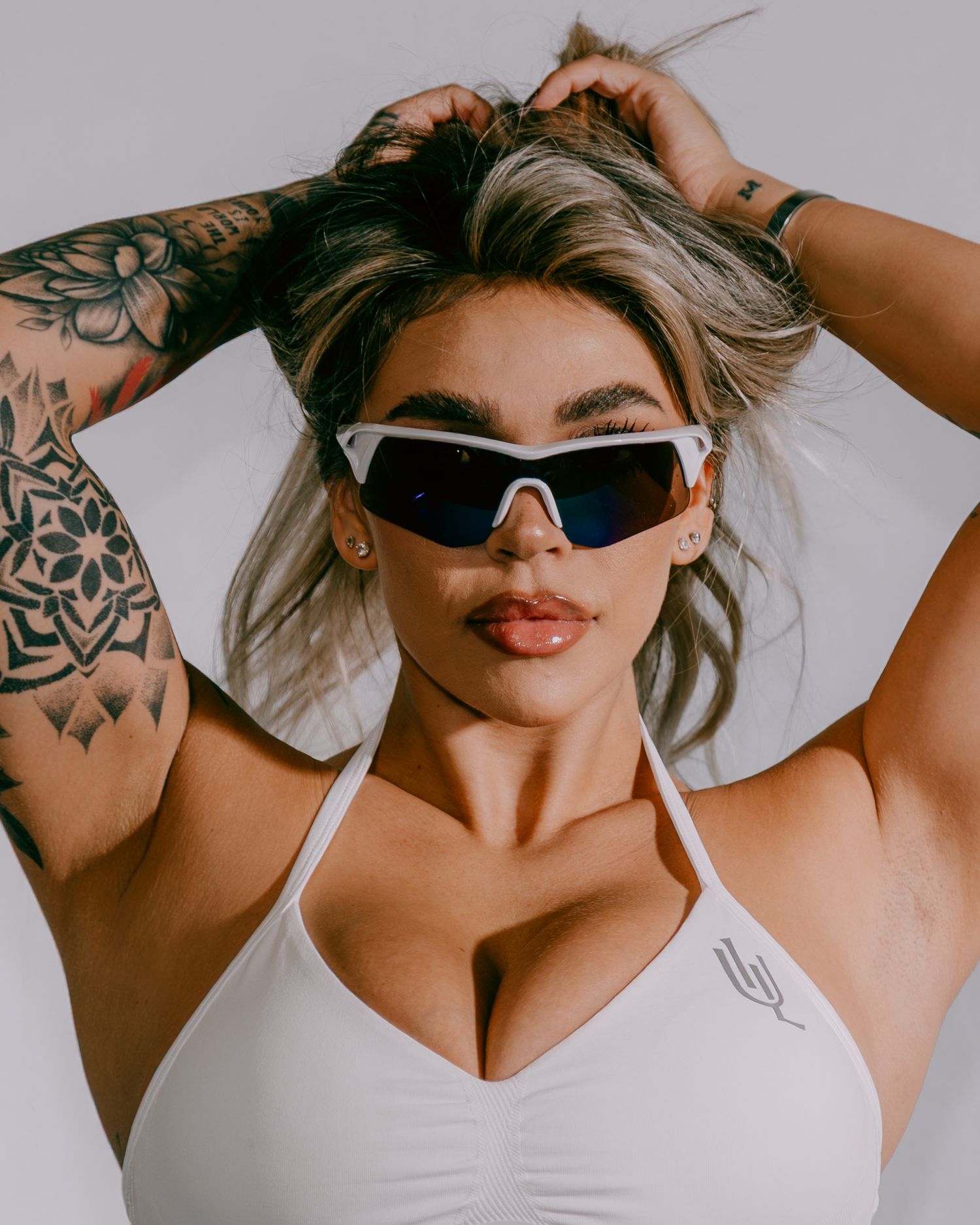 Tattooed woman in white halter top wearing futuristic white shield sunglasses, hands in blonde hair, against light background.