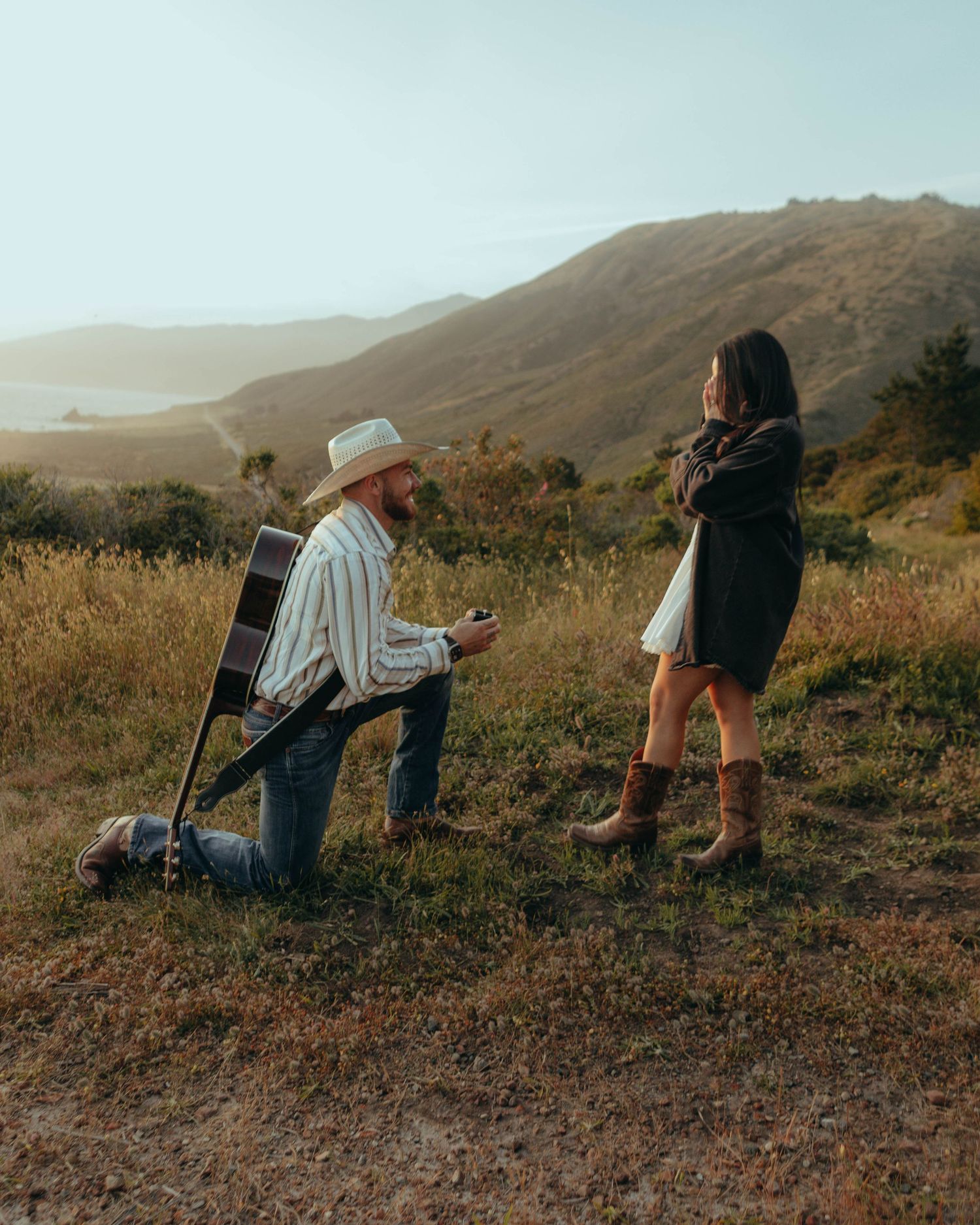 Man on one knee proposing to woman on scenic hillside with guitar and ocean views in background.