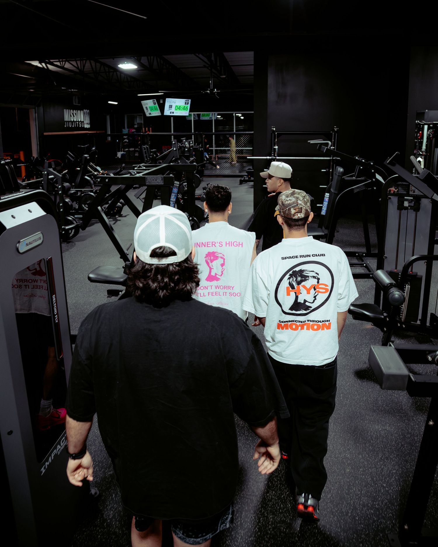 Three men in graphic tees walk through a dark gym past exercise equipment.