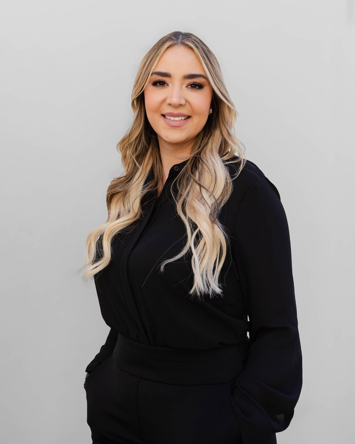 Woman in black outfit with blonde wavy hair smiling against gray background.