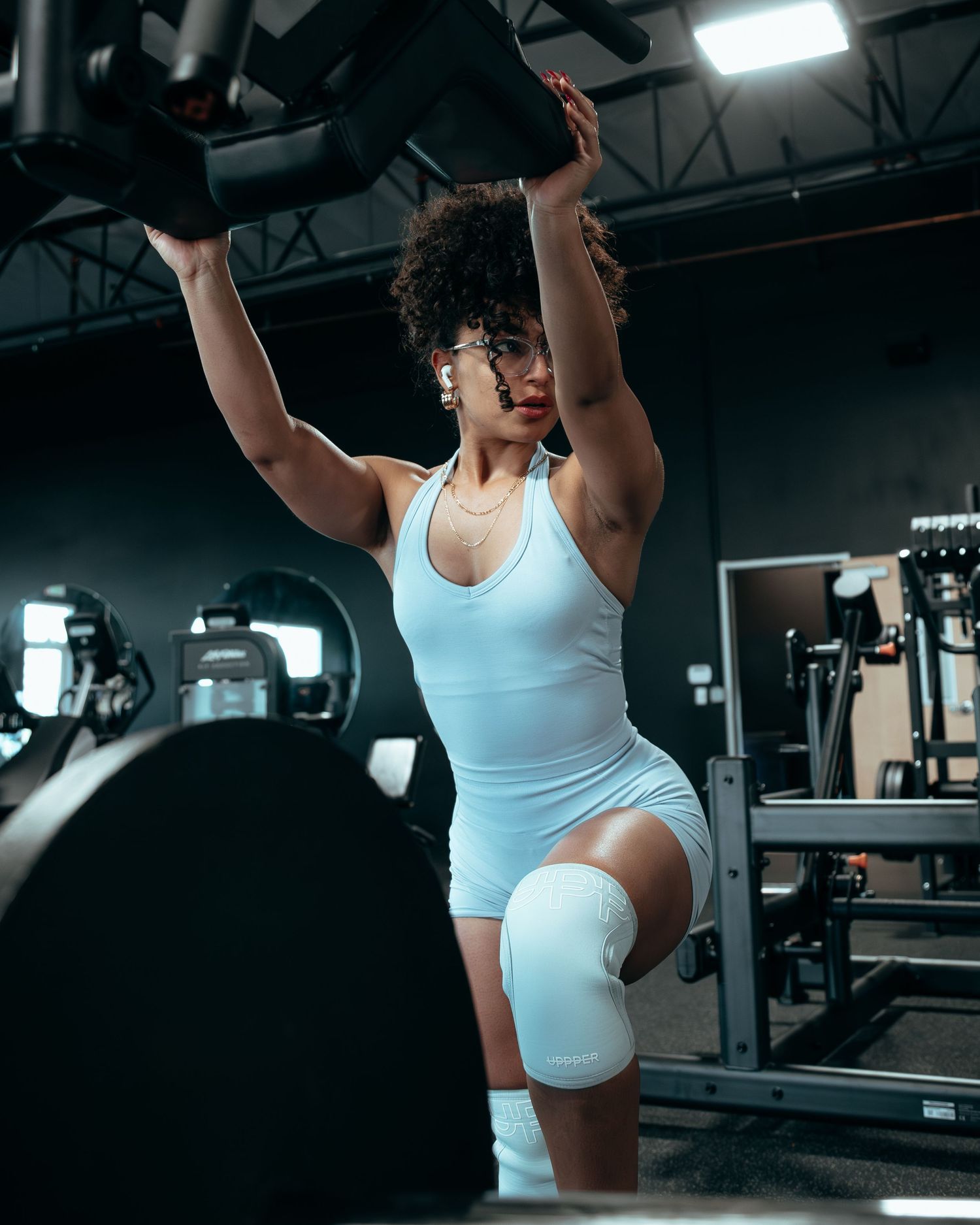 Athletic woman in white bodysuit lifting barbell overhead in dark professional gym setting.