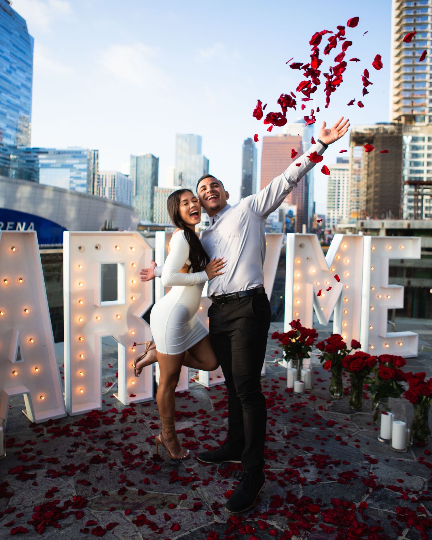 Couple celebrating proposal with rose petals and marquee marry me letters cityscape.