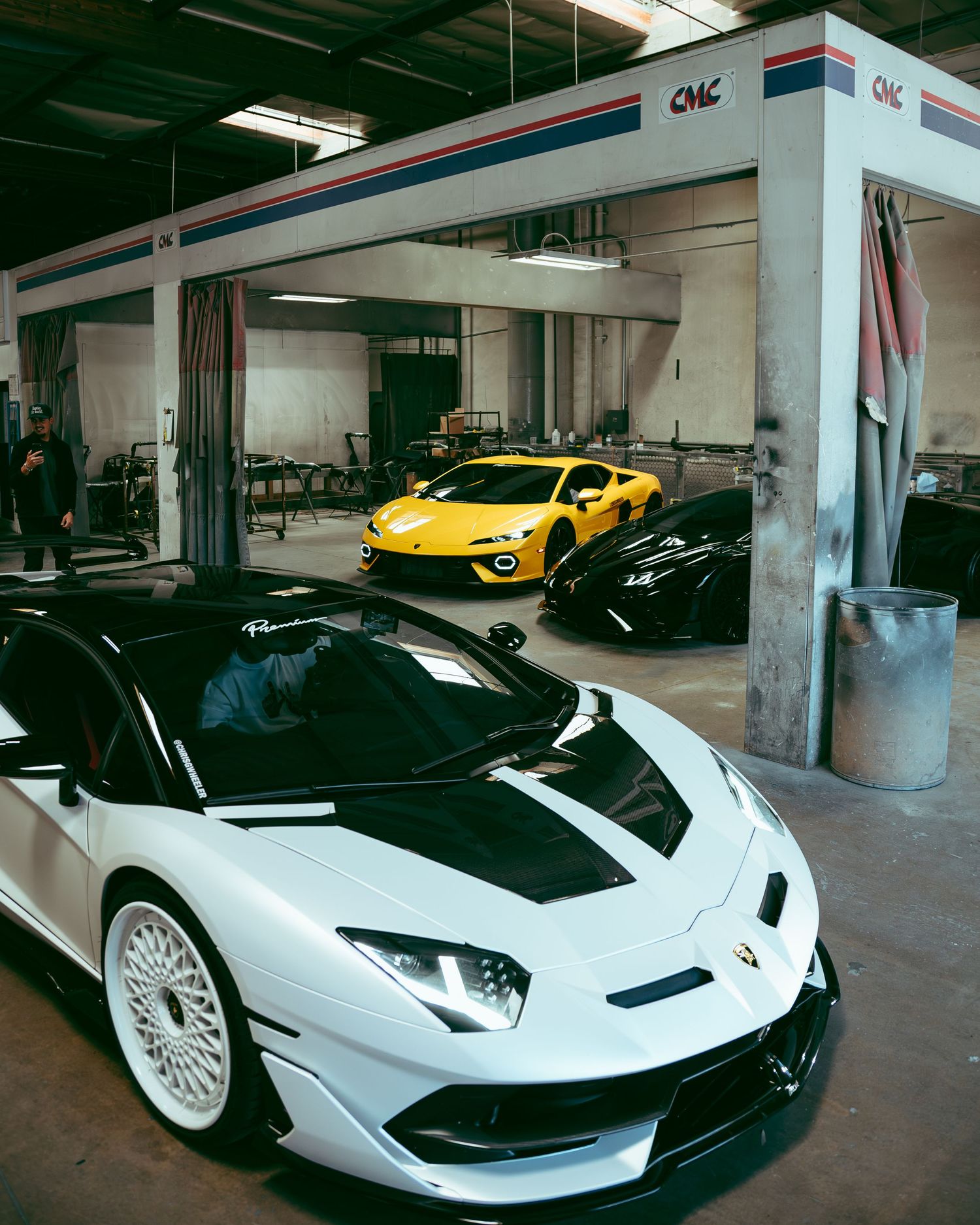 White and black Lamborghini supercar parked in front of a CMC automotive garage with yellow car inside.