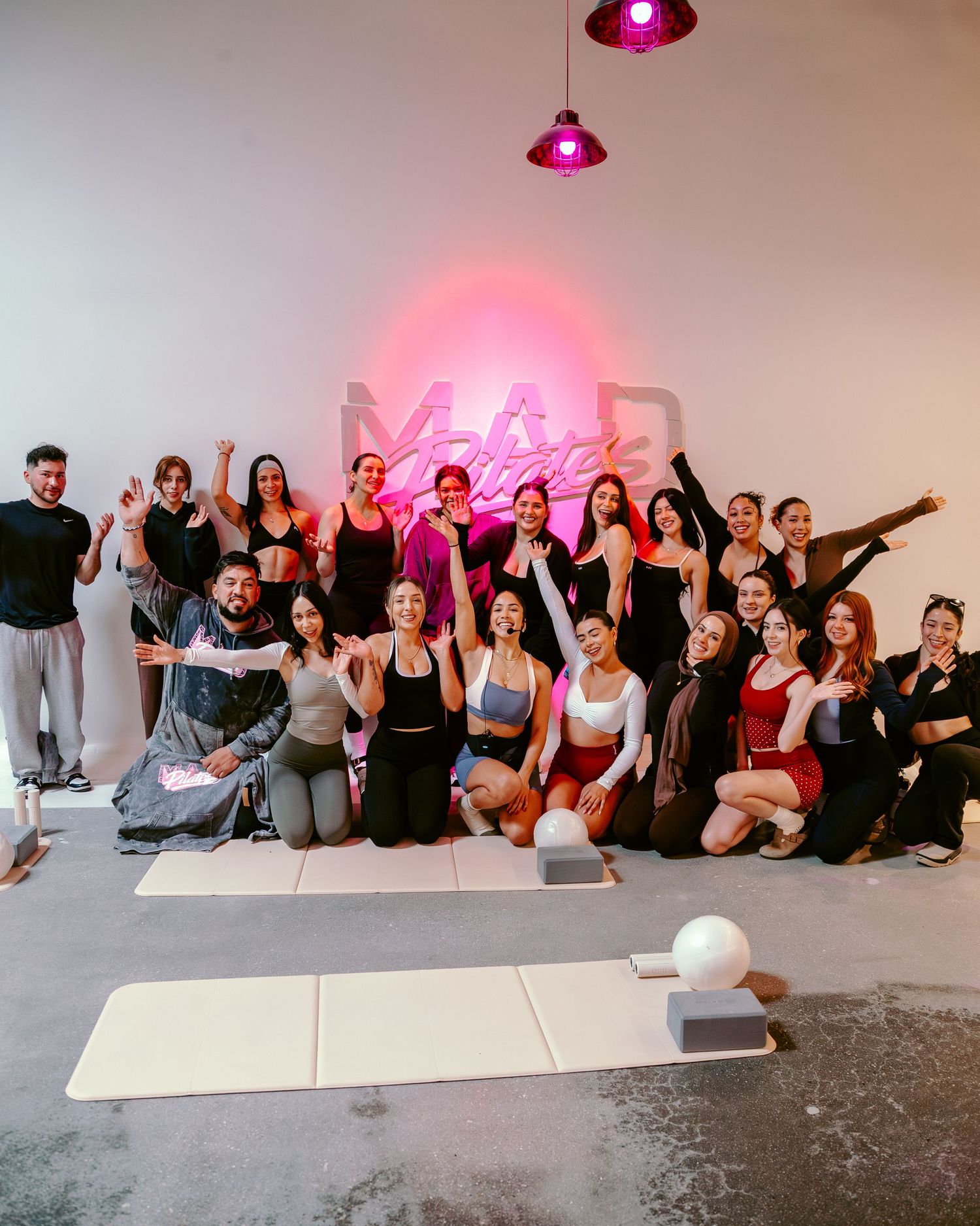 Large group poses in a yoga studio under a pink neon sign after a fitness class.